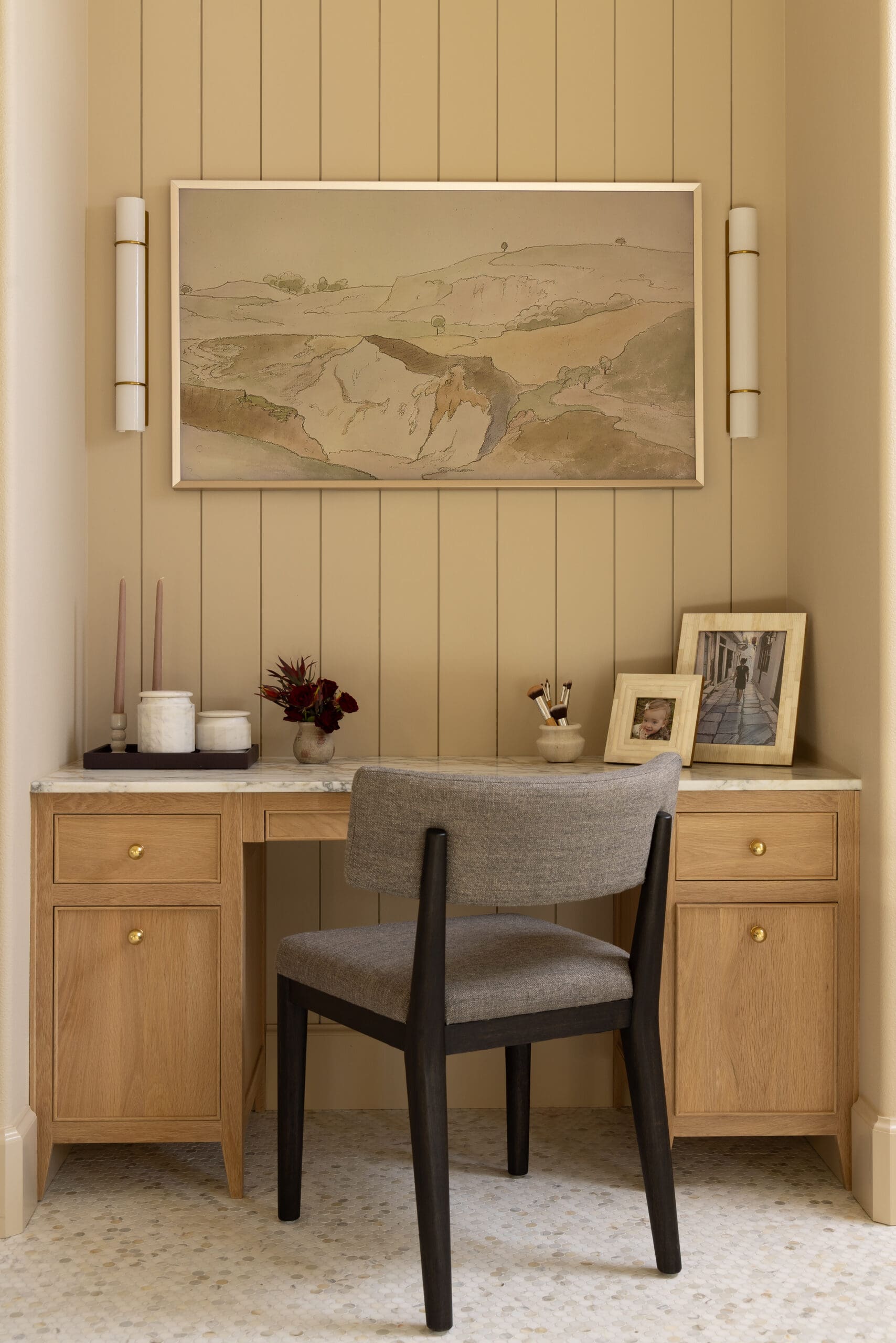 Wood desk with a gray upholstered chair in a beige paneled nook; framed landscape drawing hangs above, flanked by white sconces and personal photos on the right.