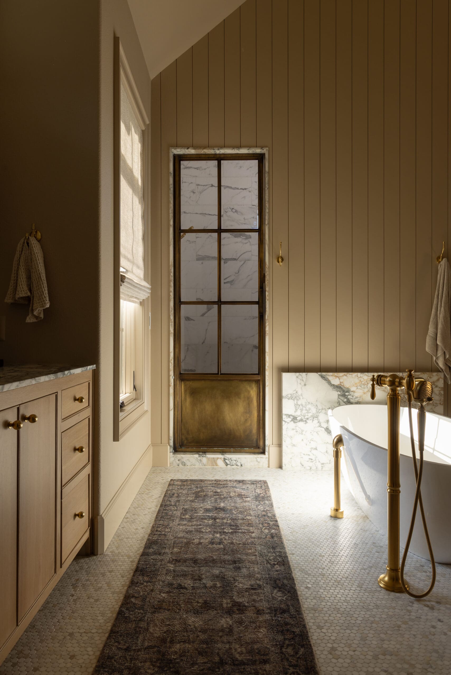 Elegant bathroom with beige vertical panel walls, a marble-framed window, and a white tub with gold fixtures.