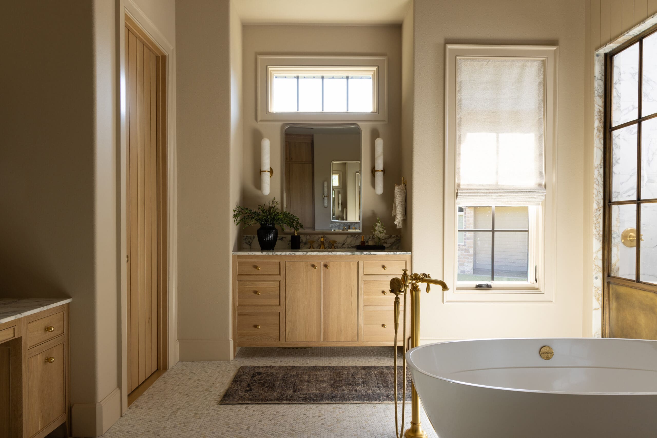 Bright luxurious bathroom with a white freestanding tub and gold faucet in the foreground, wooden vanity, and a mirror centered between wall sconces under a high window.