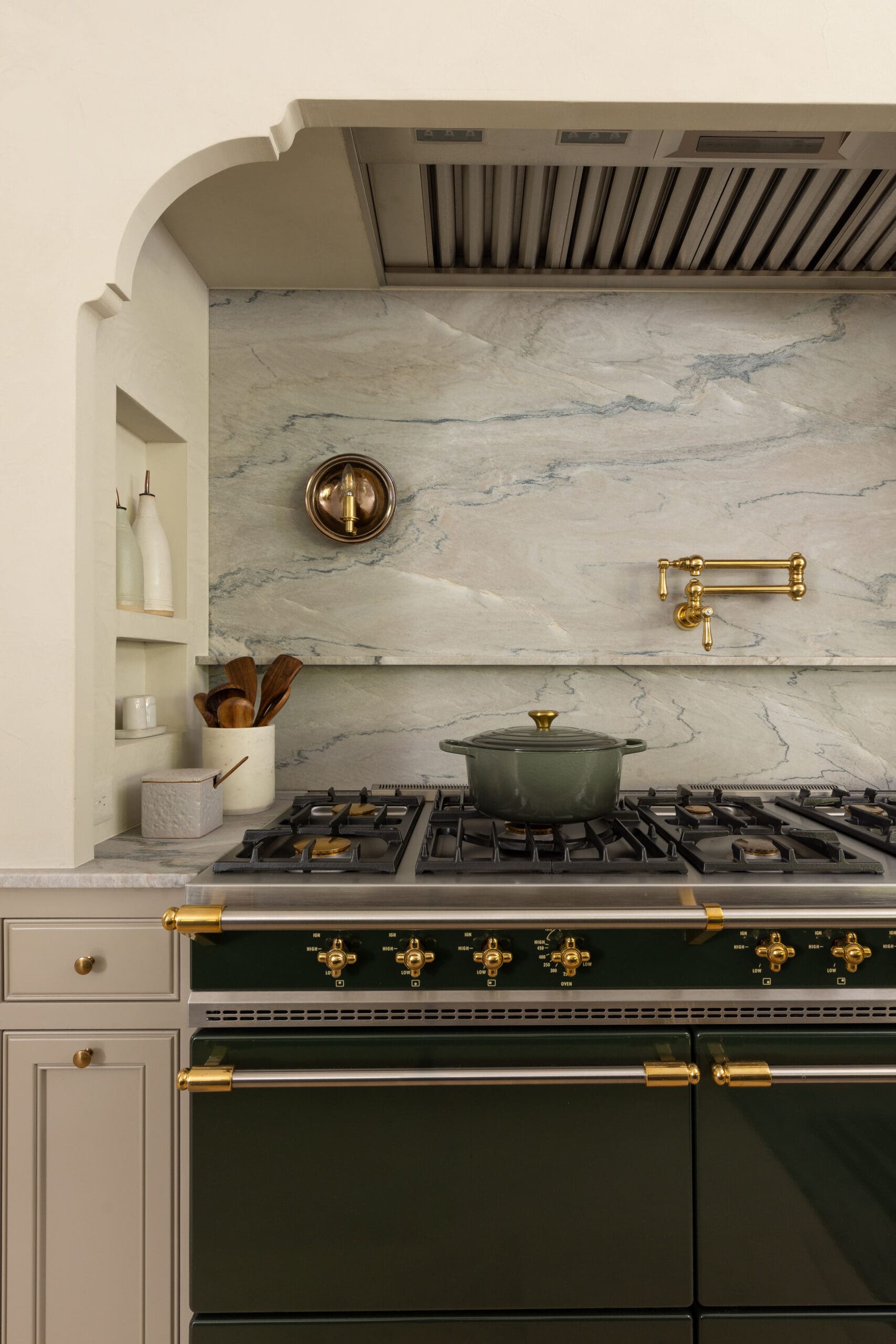 Green Dutch oven on a stainless gas range with brass handles, marble backsplash, and a range hood above.