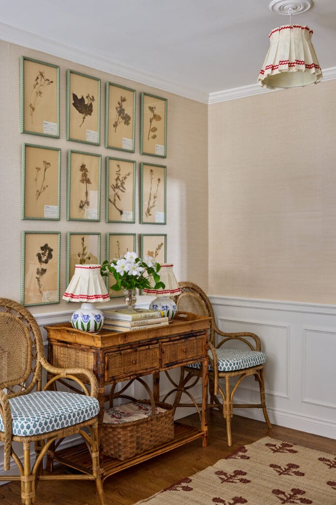 Wall features a grid of framed botanical specimens above a wicker desk with lamps and flowers; cozy vintage dining nook feel.