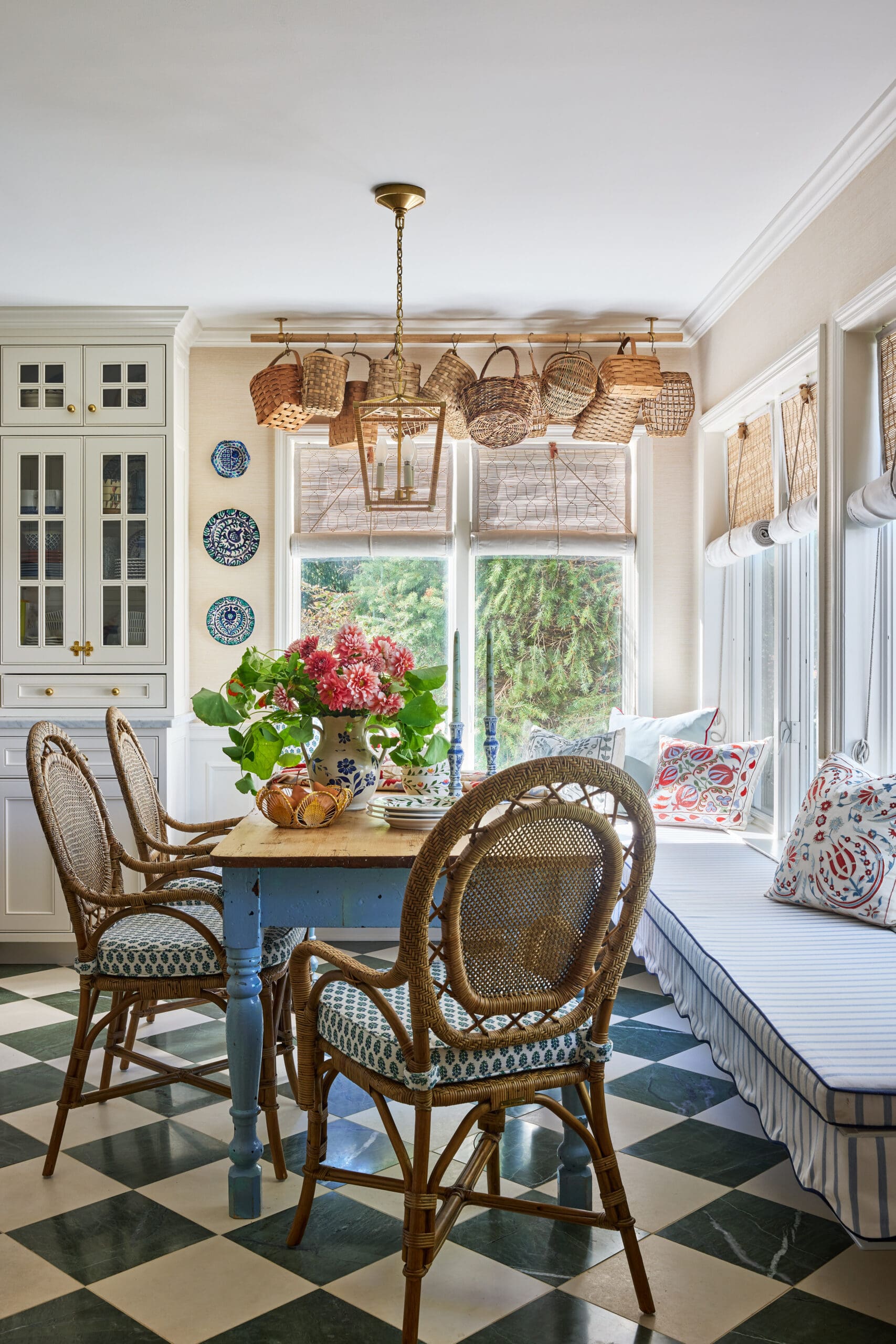 Sunlit dining area with wicker chairs around a blue-painted table, hanging baskets above, and a checkered green-and-cream floor,