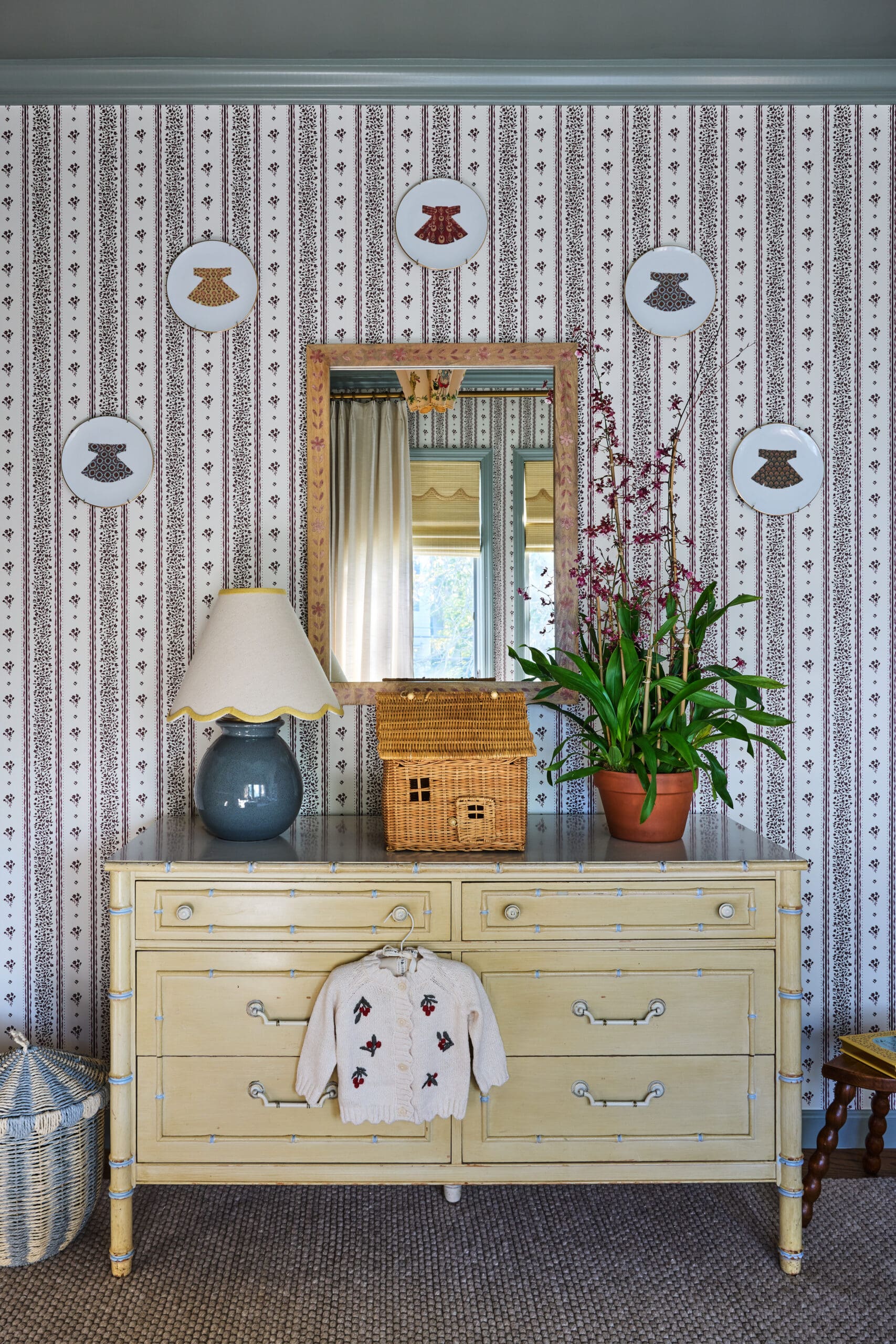 Antique pale-yellow dresser with a lamp, woven basket, and a small cardigan hanging on the front; mirror and plant against striped wallpaper.