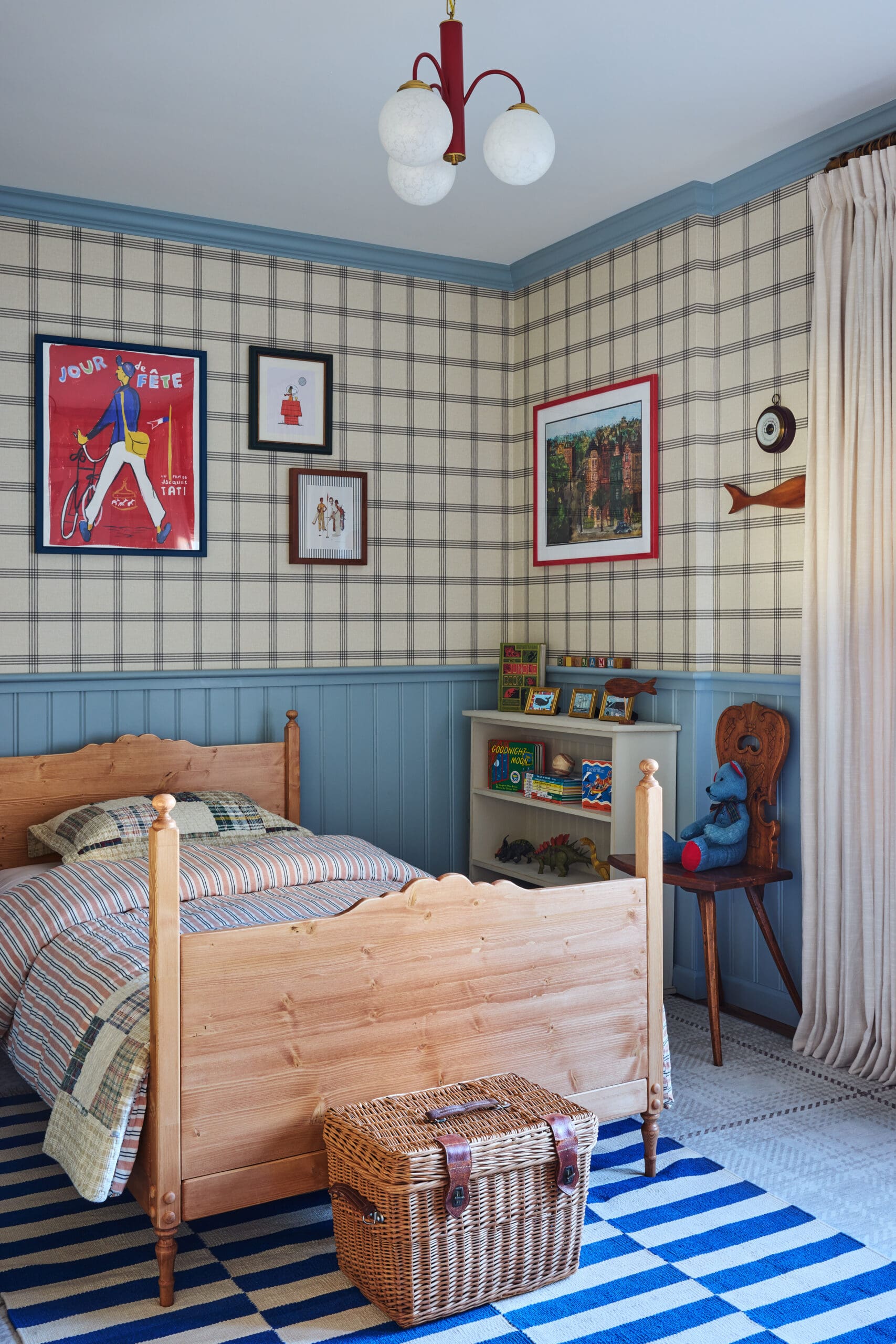 Child’s bedroom with a wooden bed, blue trim, plaid wallpaper, framed art, and a wicker basket on a striped rug cabined by a light-filled corner.