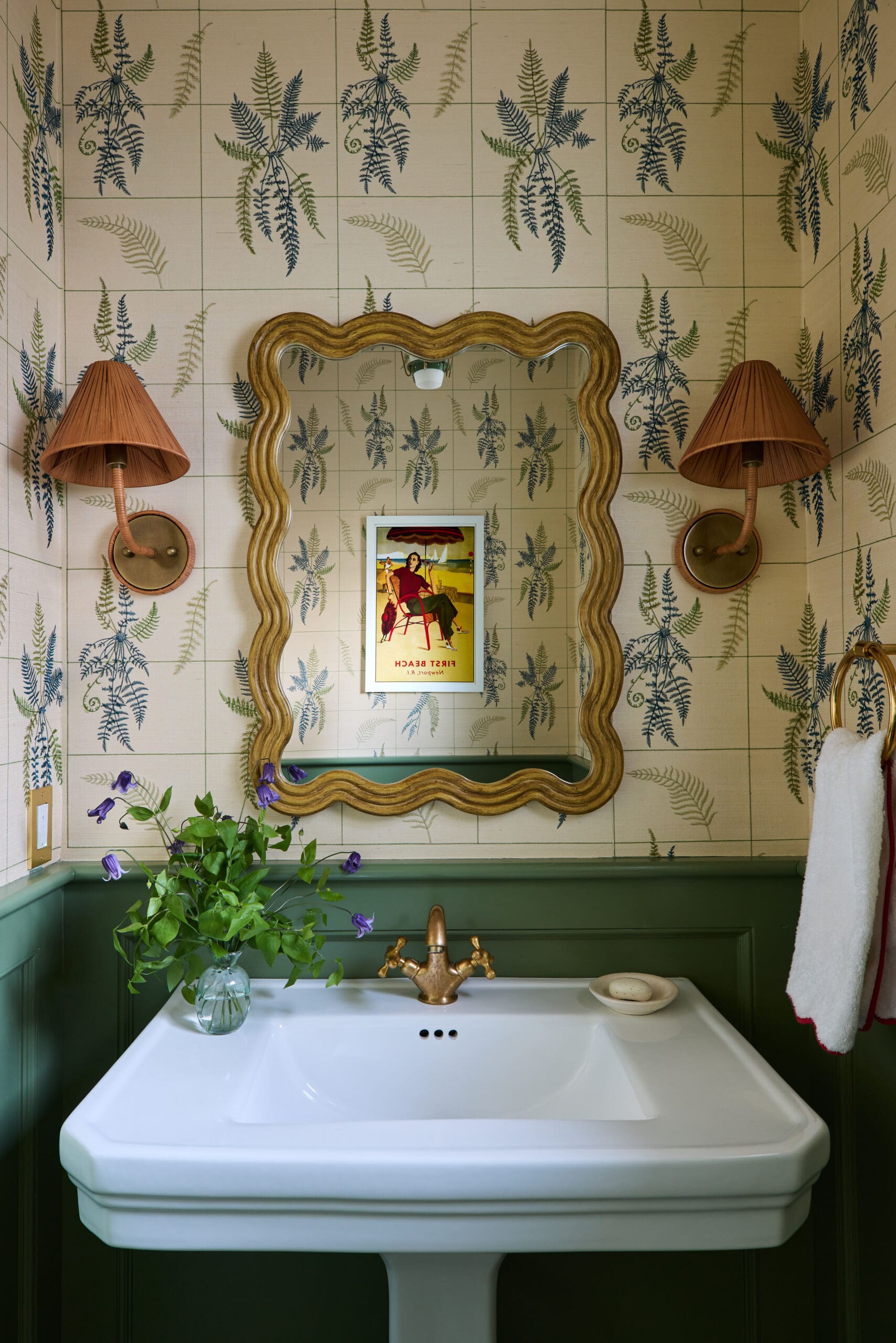 Bathroom scene with a scalloped gold mirror, orange lamps, fern-pattern wallpaper, a white pedestal sink, and a vase of purple flowers.