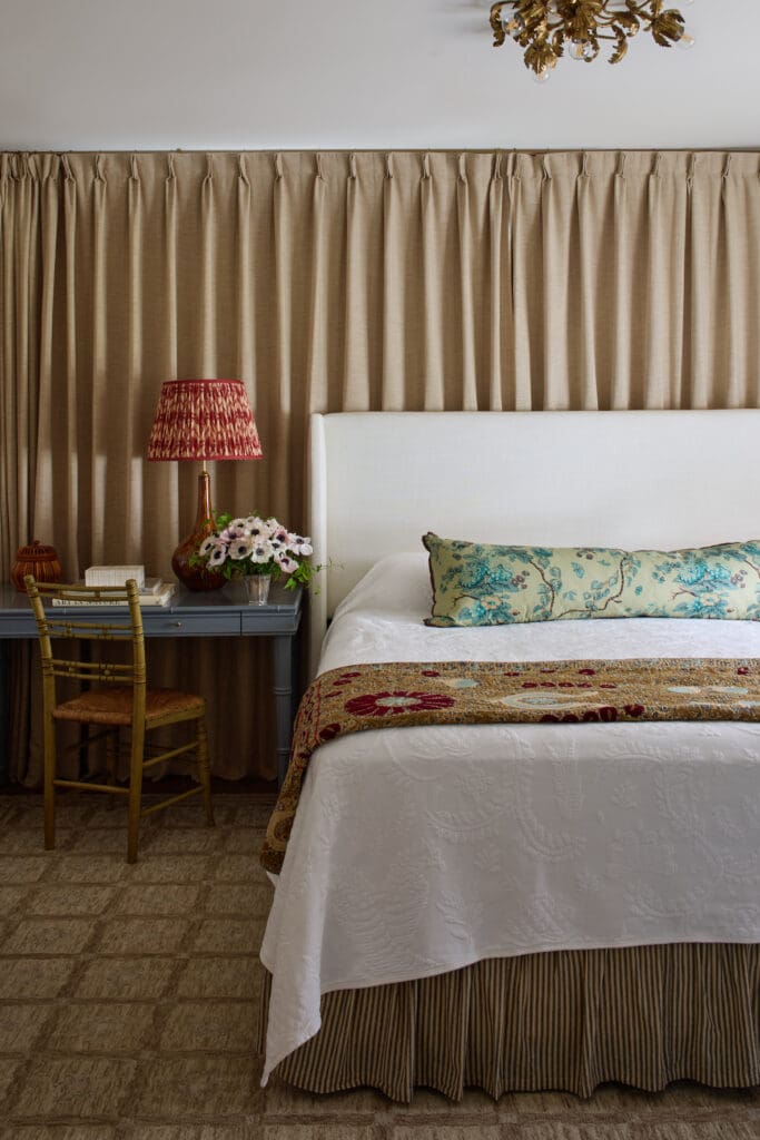 Hotel-style bedroom with a white bed, beige curtains, and a red patterned lamp on a gray nightstand with flowers nearby.