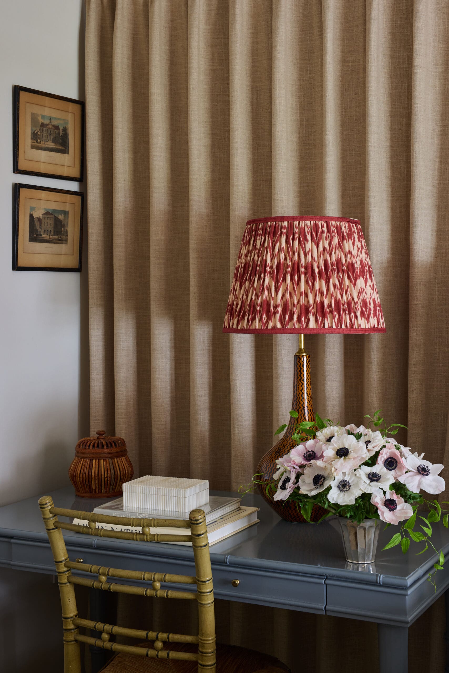 Lamp with red patterned shade on a glass-topped table beside a bouquet of white and pink flowers, beige drapes in the background.