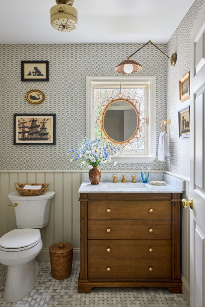 Traditional bathroom with a white toilet beside a tall wooden dresser vanity, brass hardware, and a round wicker-motif mirror above a marble sink area; blue-toned patterned wallpaper and a vase of blue and white flowers add color.