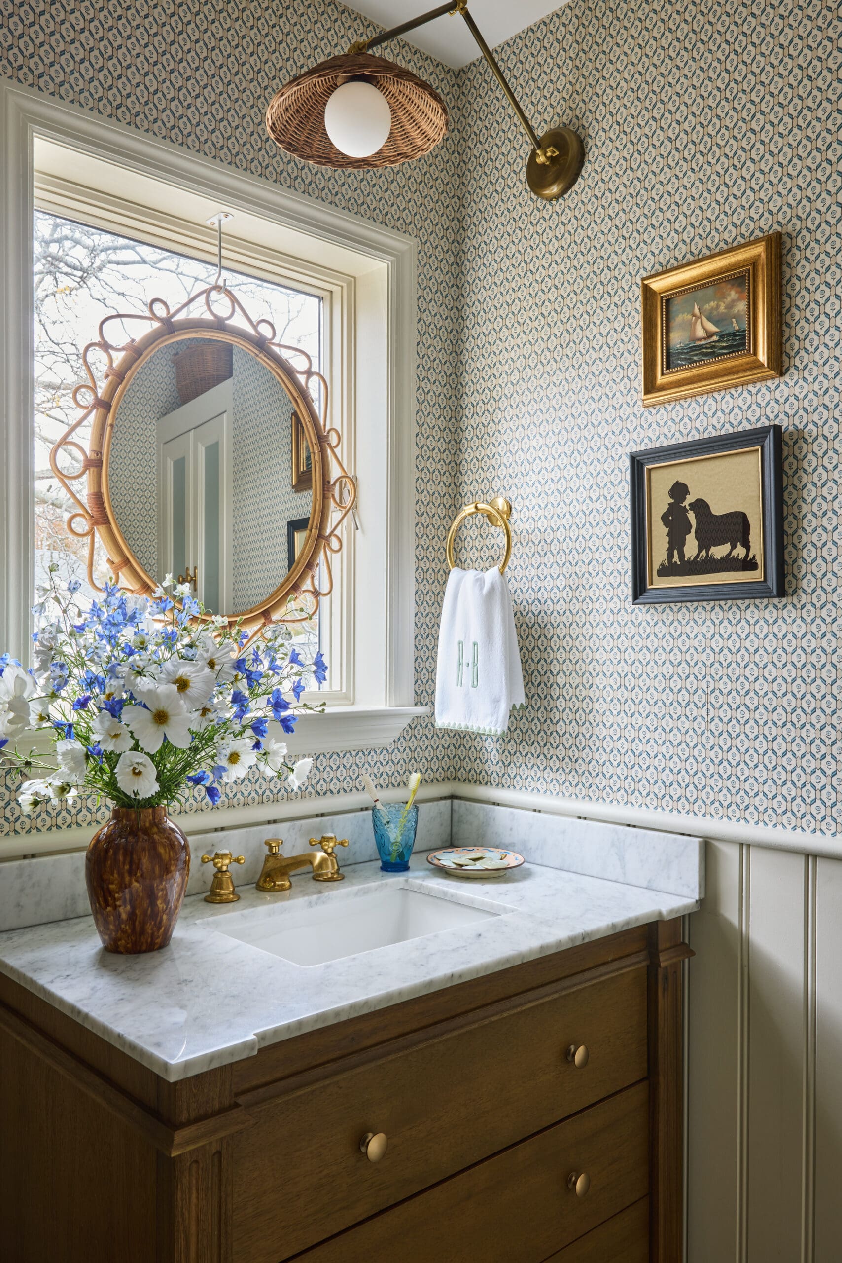 Wood vanity with marble countertop, wicker-framed round mirror, and a vase of white and blue flowers in a blue patterned bathroom wall.