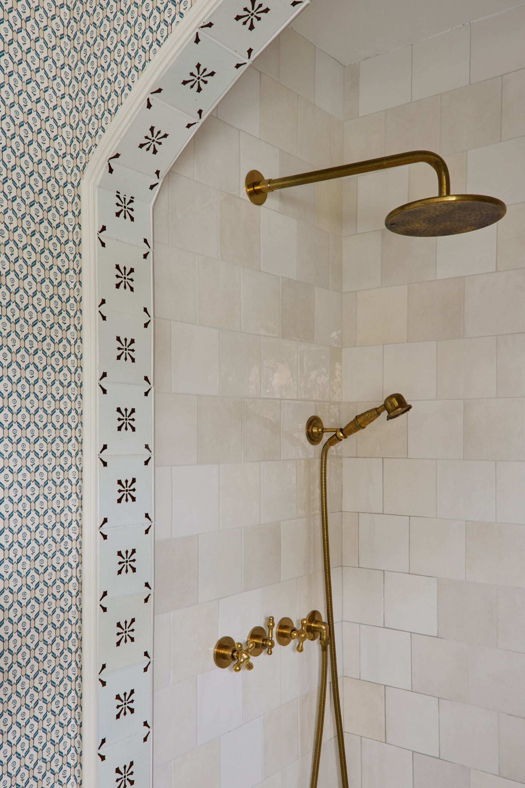 Brass showerhead and faucet with handheld hose mounted on white tiled shower wall, bordered by decorative white tiles with brown star patterns on the left.