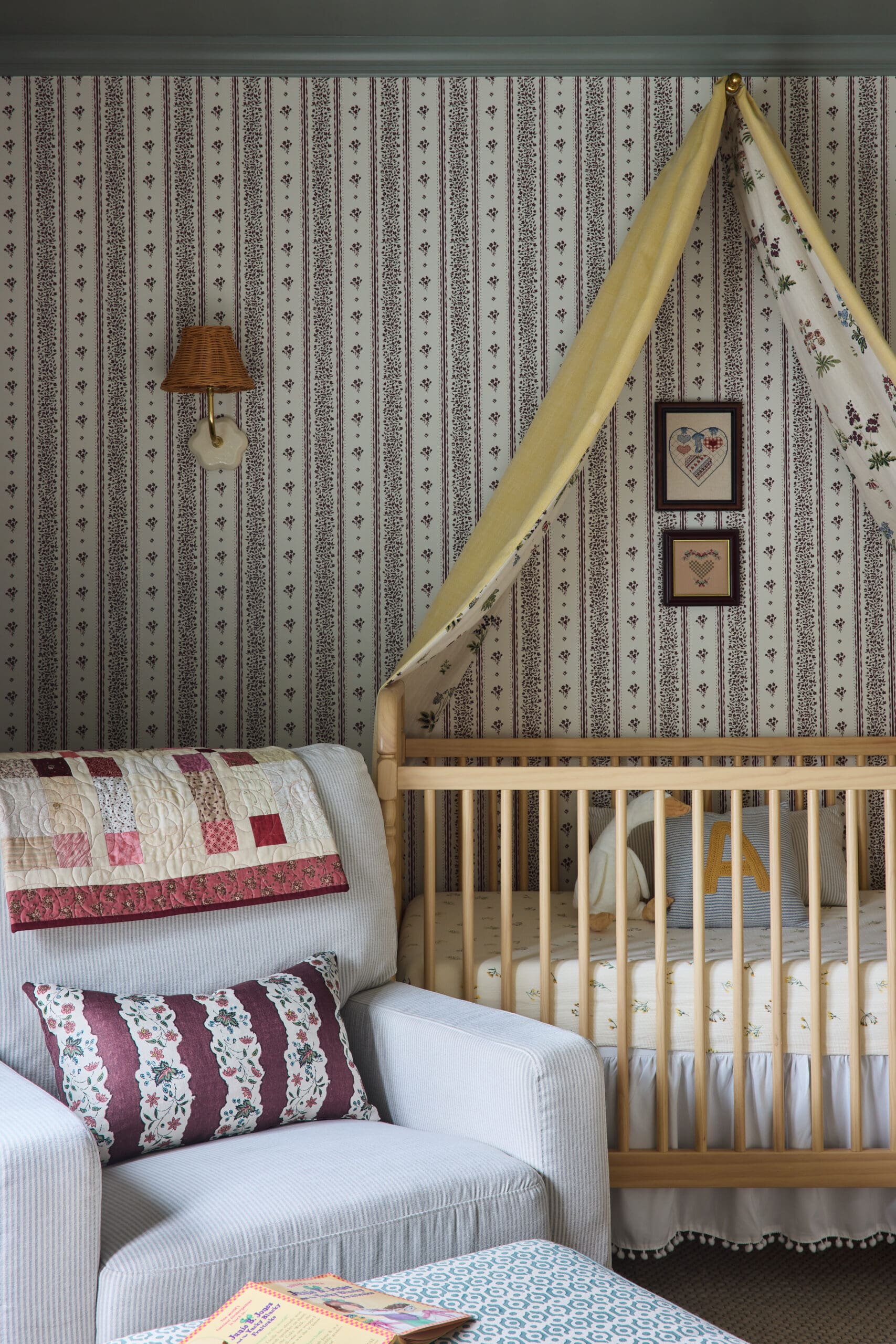 Nursery corner with a wooden crib, yellow canopy, burgundy striped wallpaper, and framed heart art.