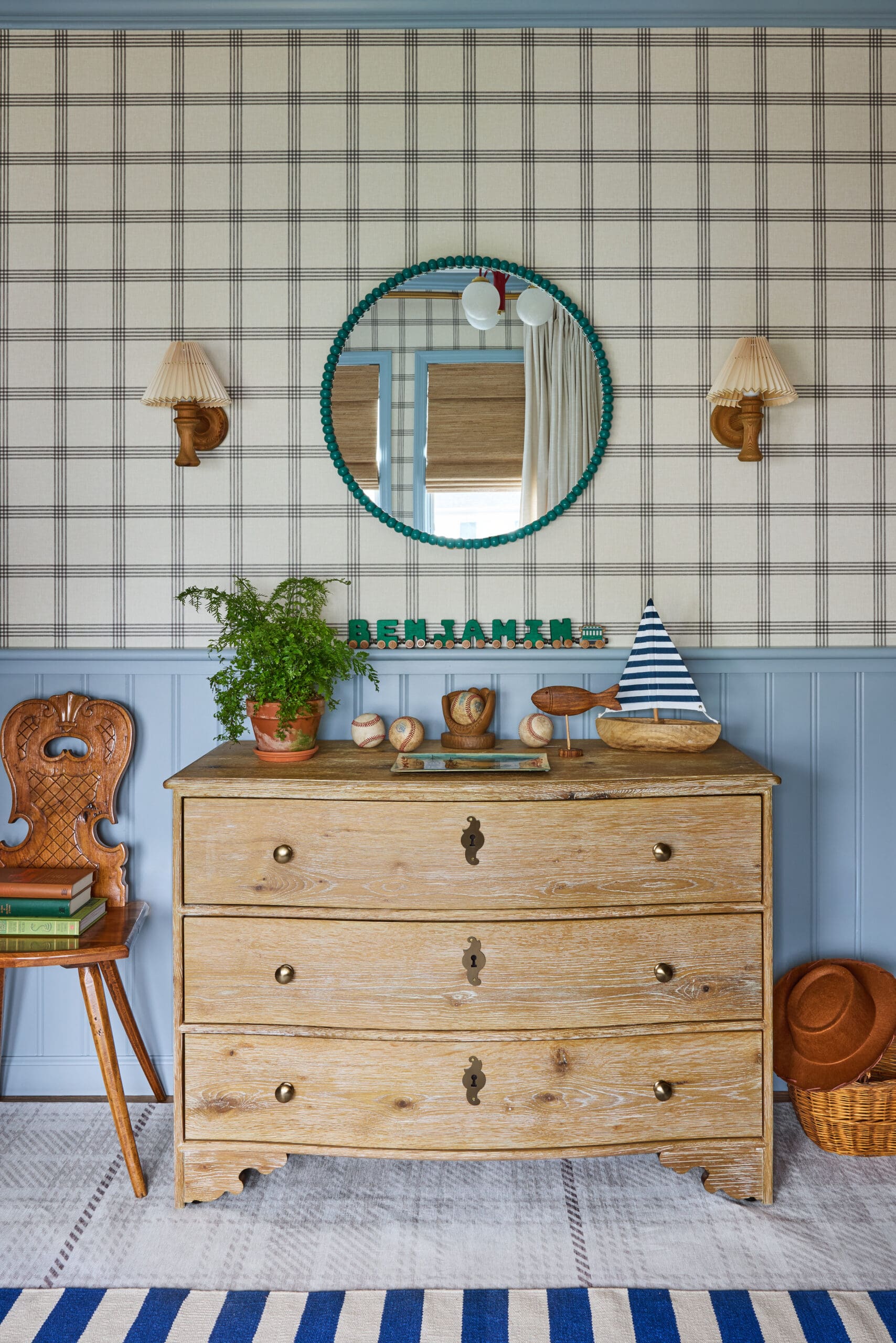Vintage wooden dresser with a teal round mirror, plaid wallpaper, and coastal decor including a plant and toy boat.