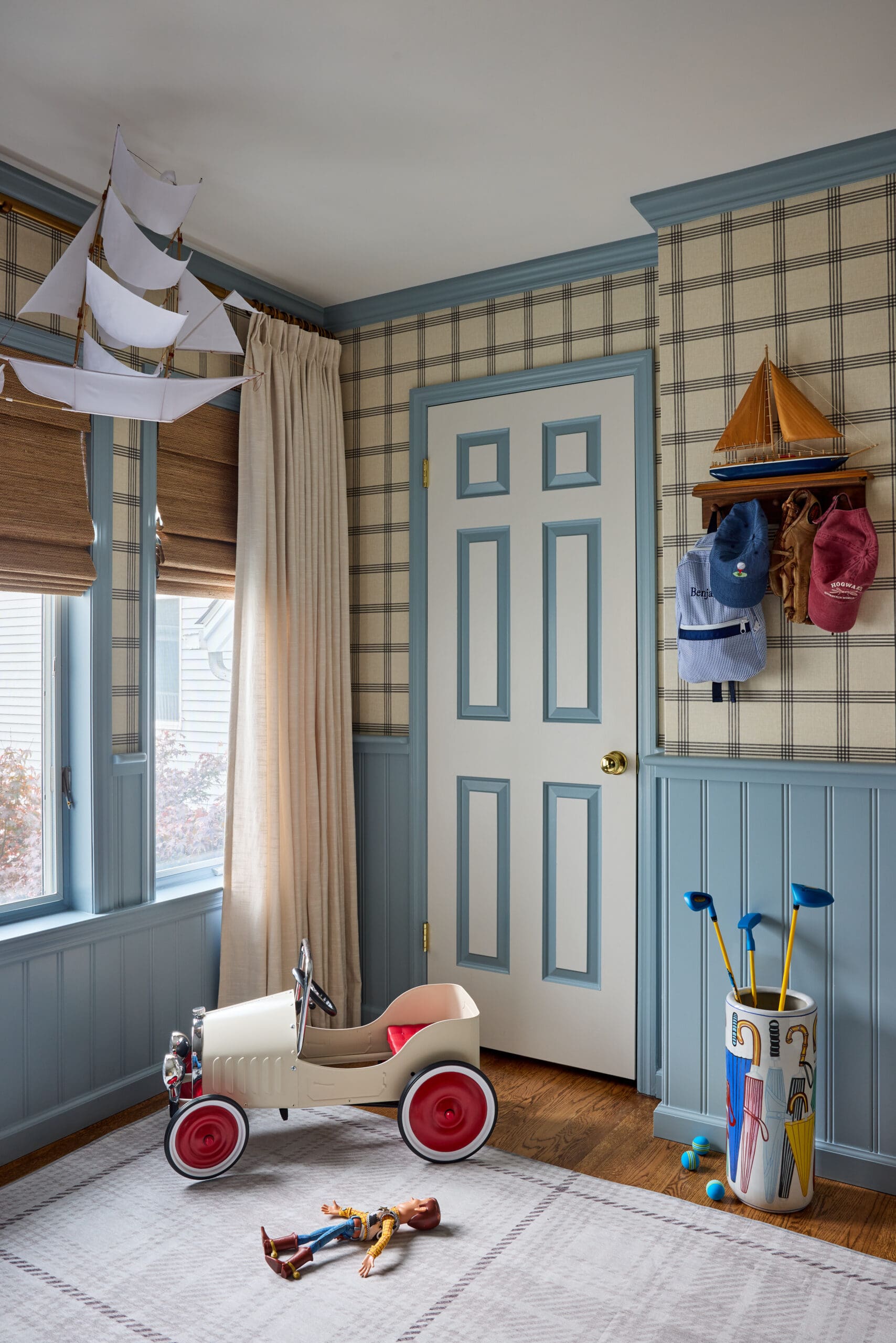 Child's playroom corner with a white toy sailboat hanging from the ceiling and a pale blue-trimmed door, plaid wallpaper, and hats on a wall hook.