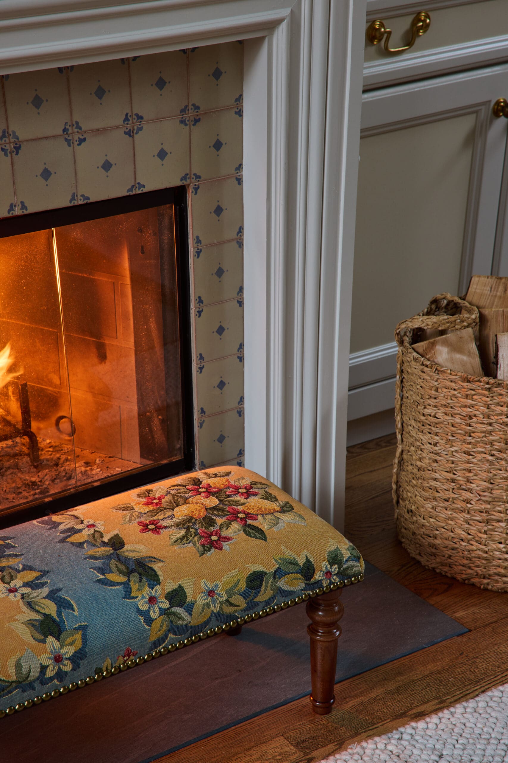 Fireplace with beige tiled surround featuring blue pattern, glass door glowing with a fire, beside a floral upholstered bench and wicker basket of firewood.