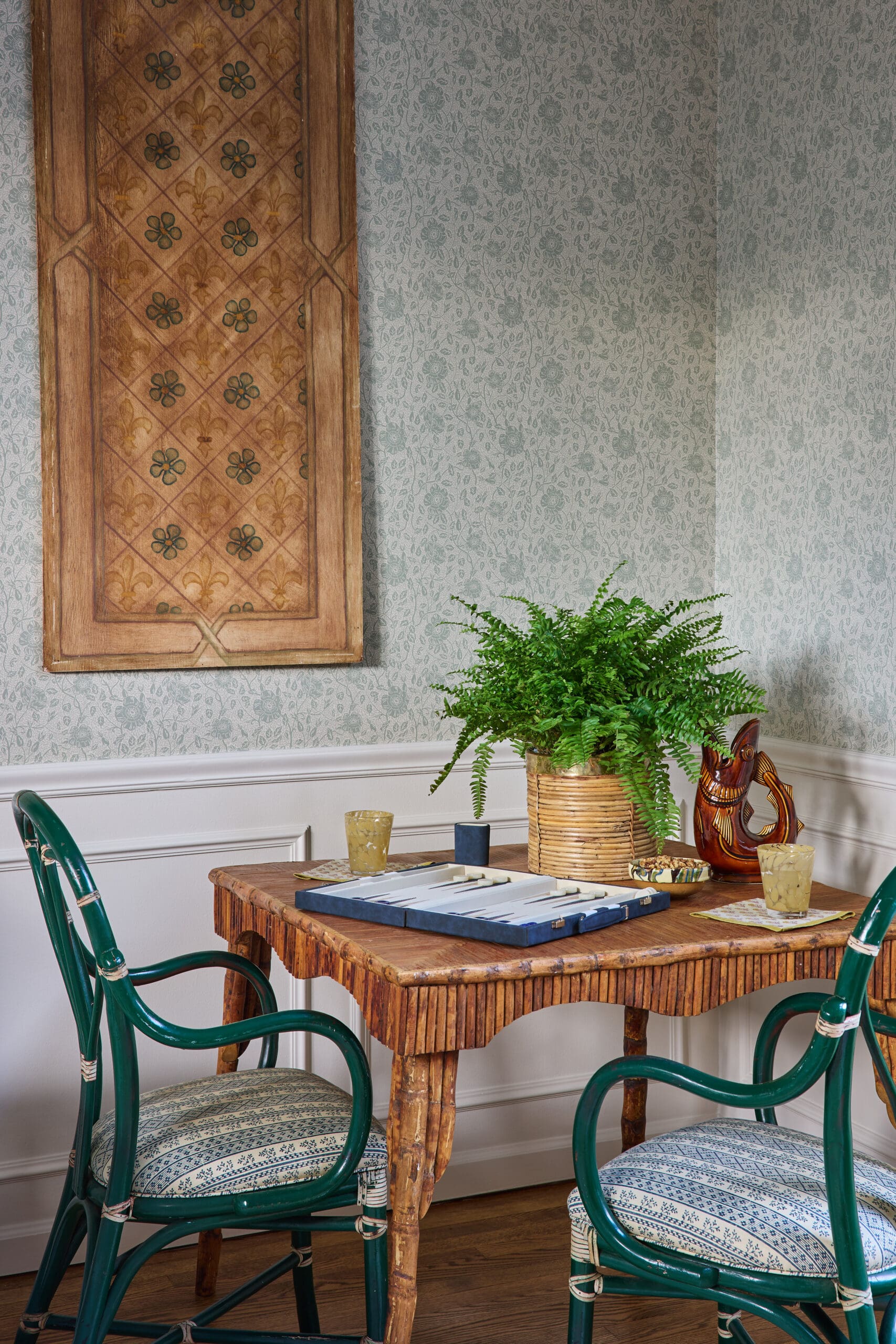 Wooden dining table with green chairs, a potted fern in a basket, and a vintage carved cabinet against floral wallpaper.