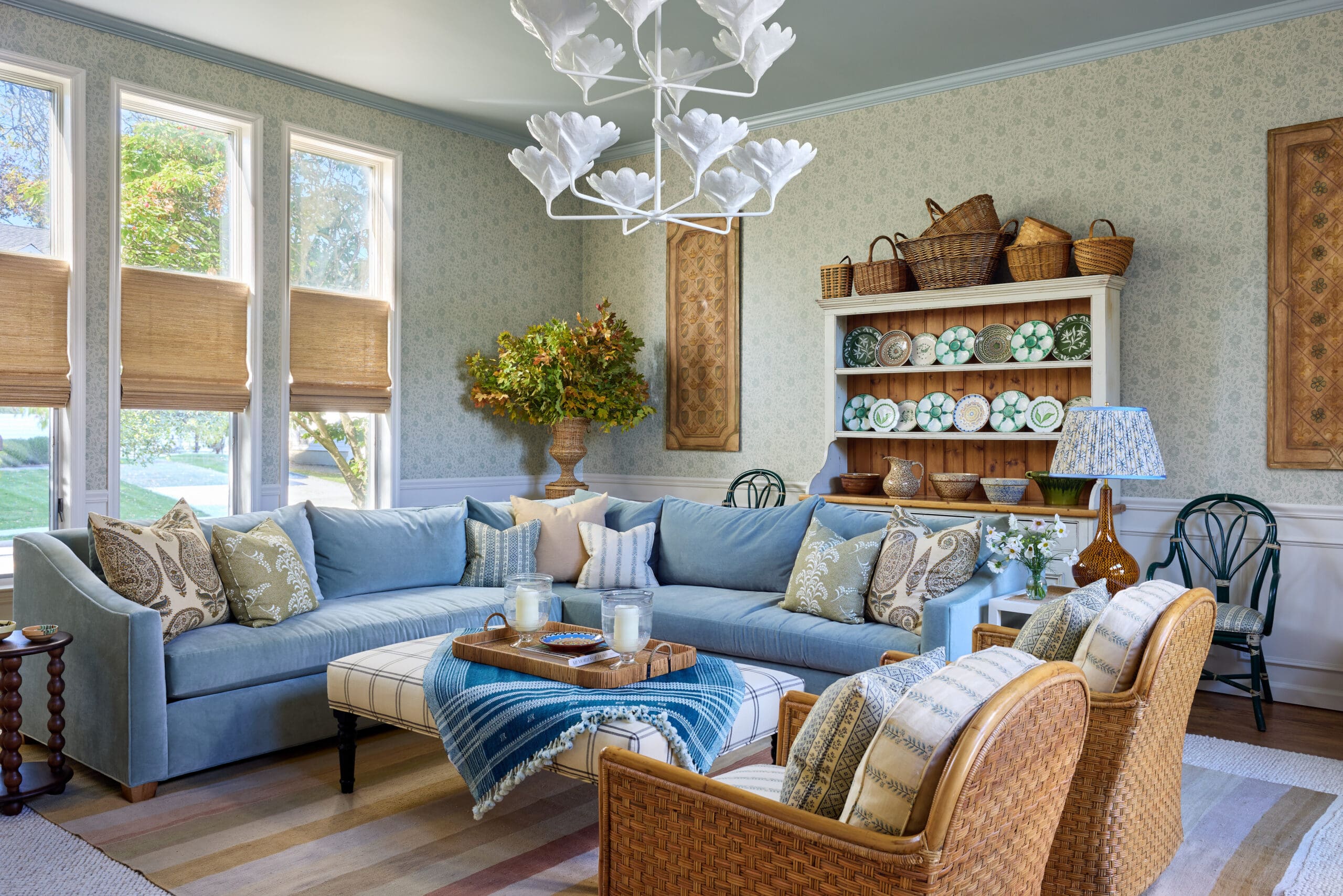 Bright living room with a blue sectional, patterned pillows, and wicker chairs around a tray table under a white chandelier.