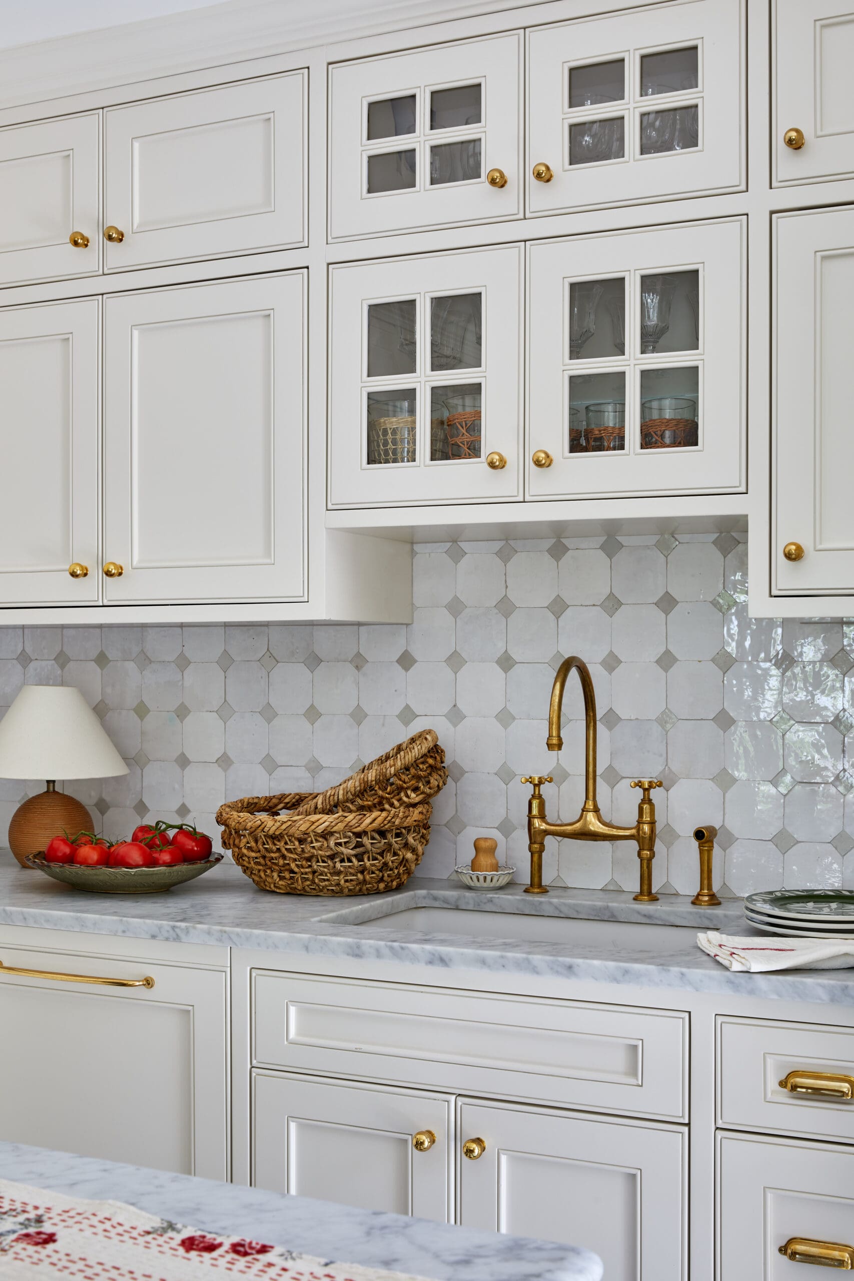 White kitchen with matching cabinets, glass-front doors, and brass hardware; marble countertop and bowls of tomatoes in view.