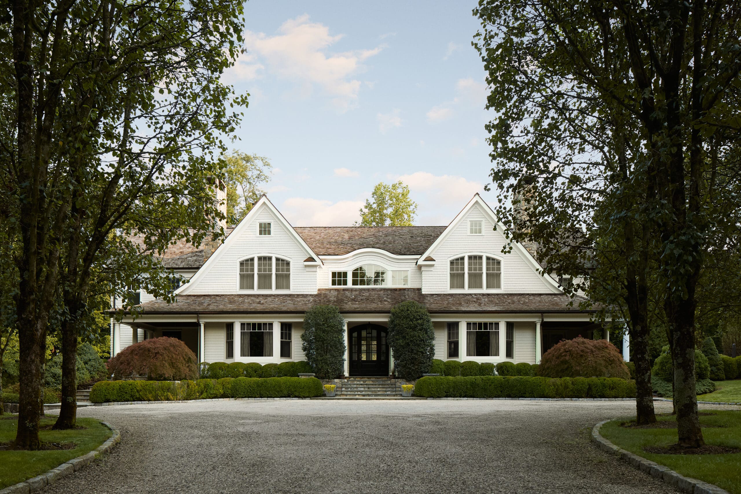 White two-story house with arched dormers framed by green trees on a gravel driveway, front garden visible down the path.