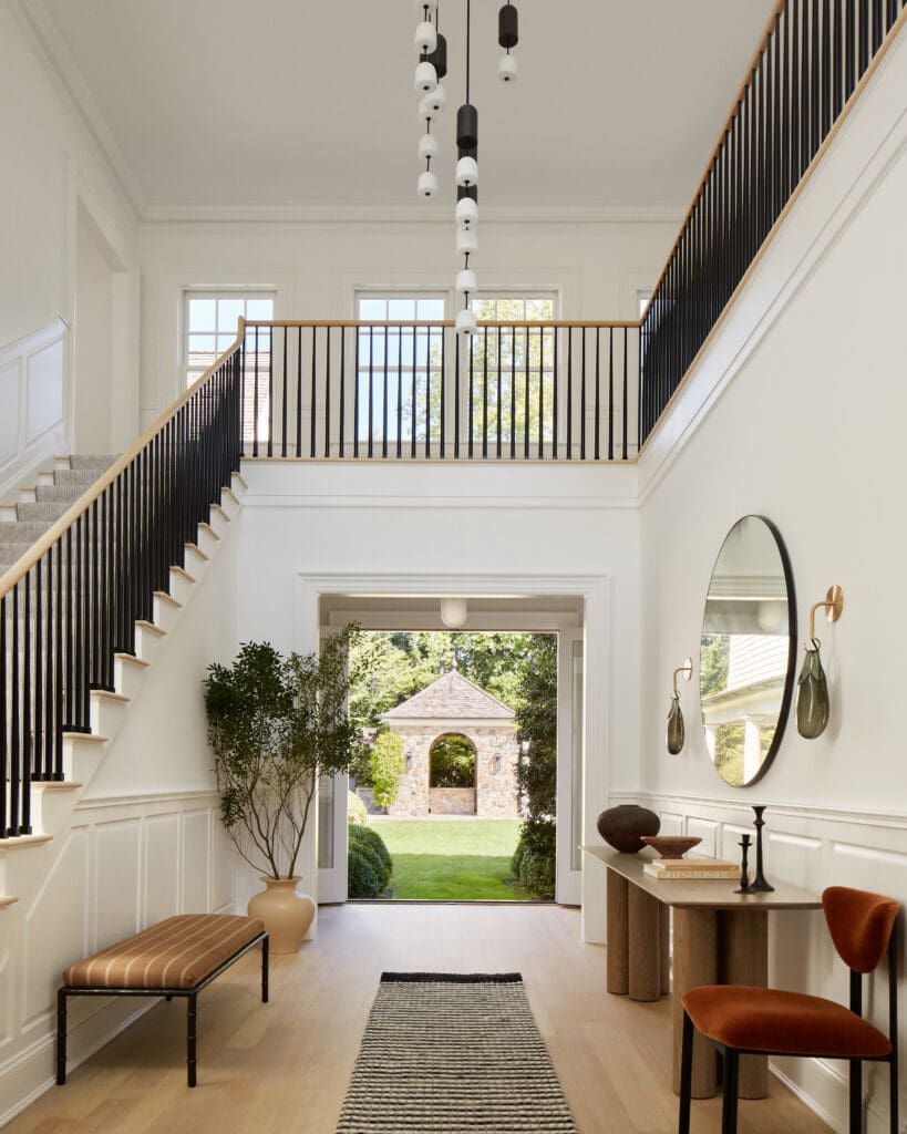 Bright entry hall with two sweeping staircases and black iron balusters meeting at a balcony, opening to a sunny garden courtyard.