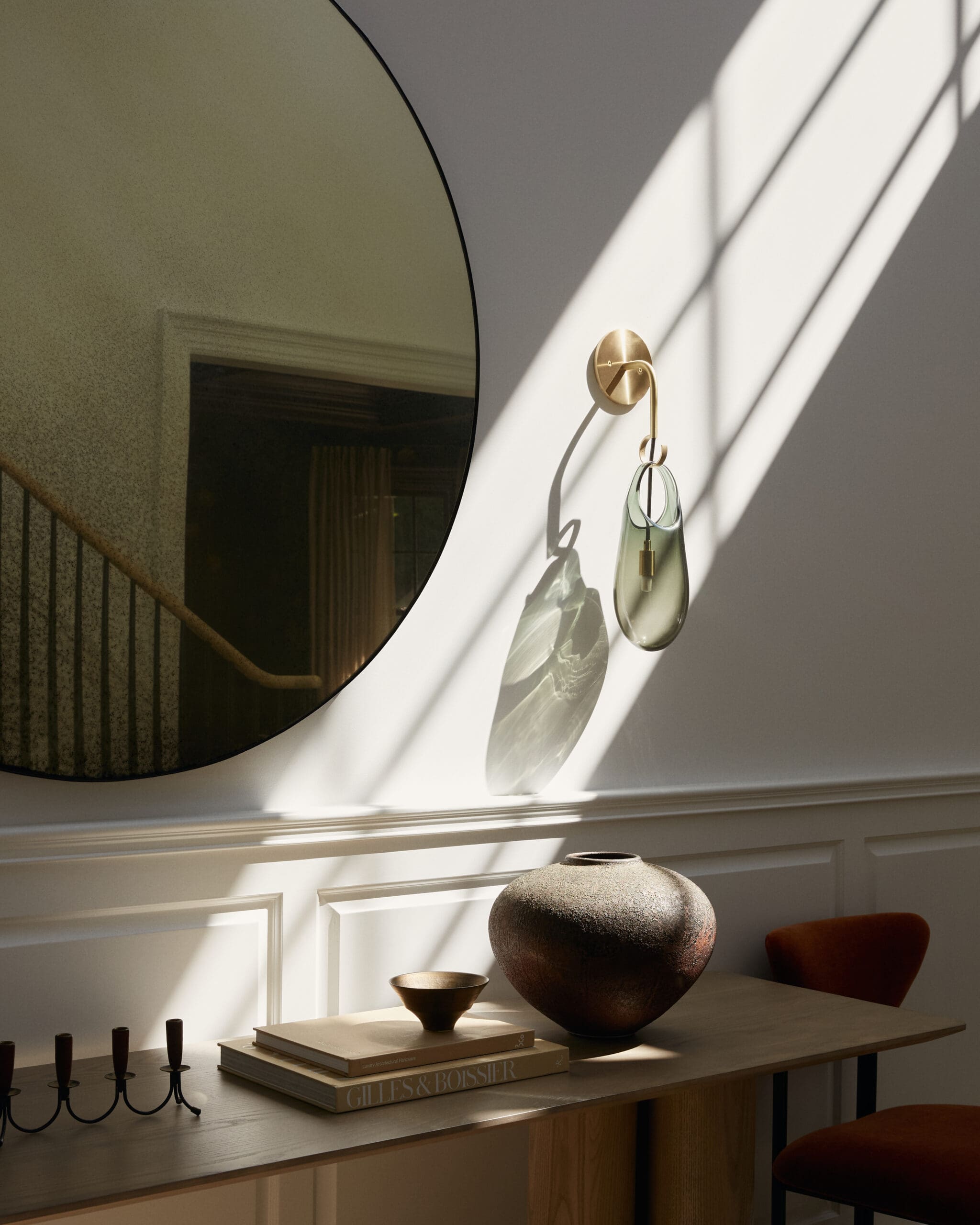 Round wall mirror reflecting a staircase and interior, with a brass wall hook and glass hanging ornament catching light.