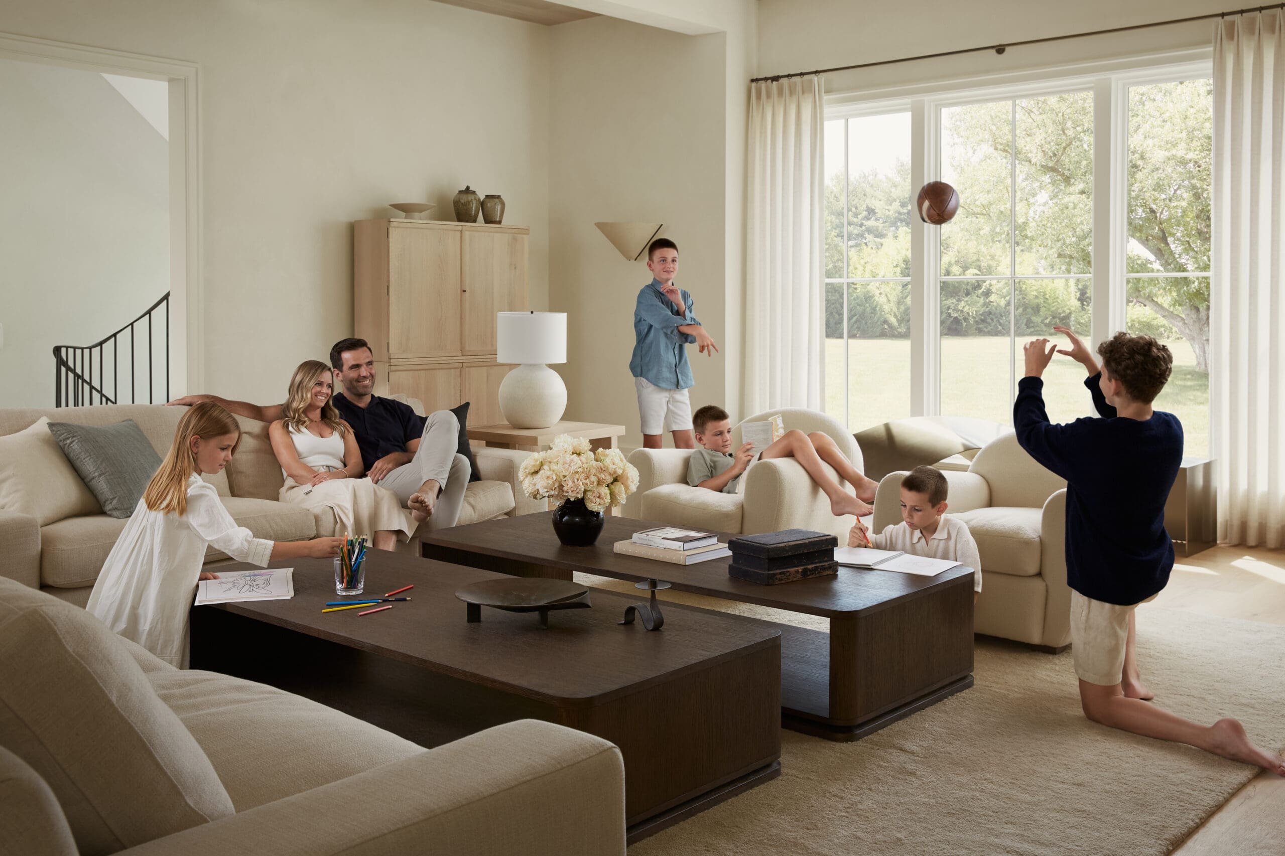 Family living room with kids playing indoors while parents relax on a sofa; a boy throws a basketball by the large windows.