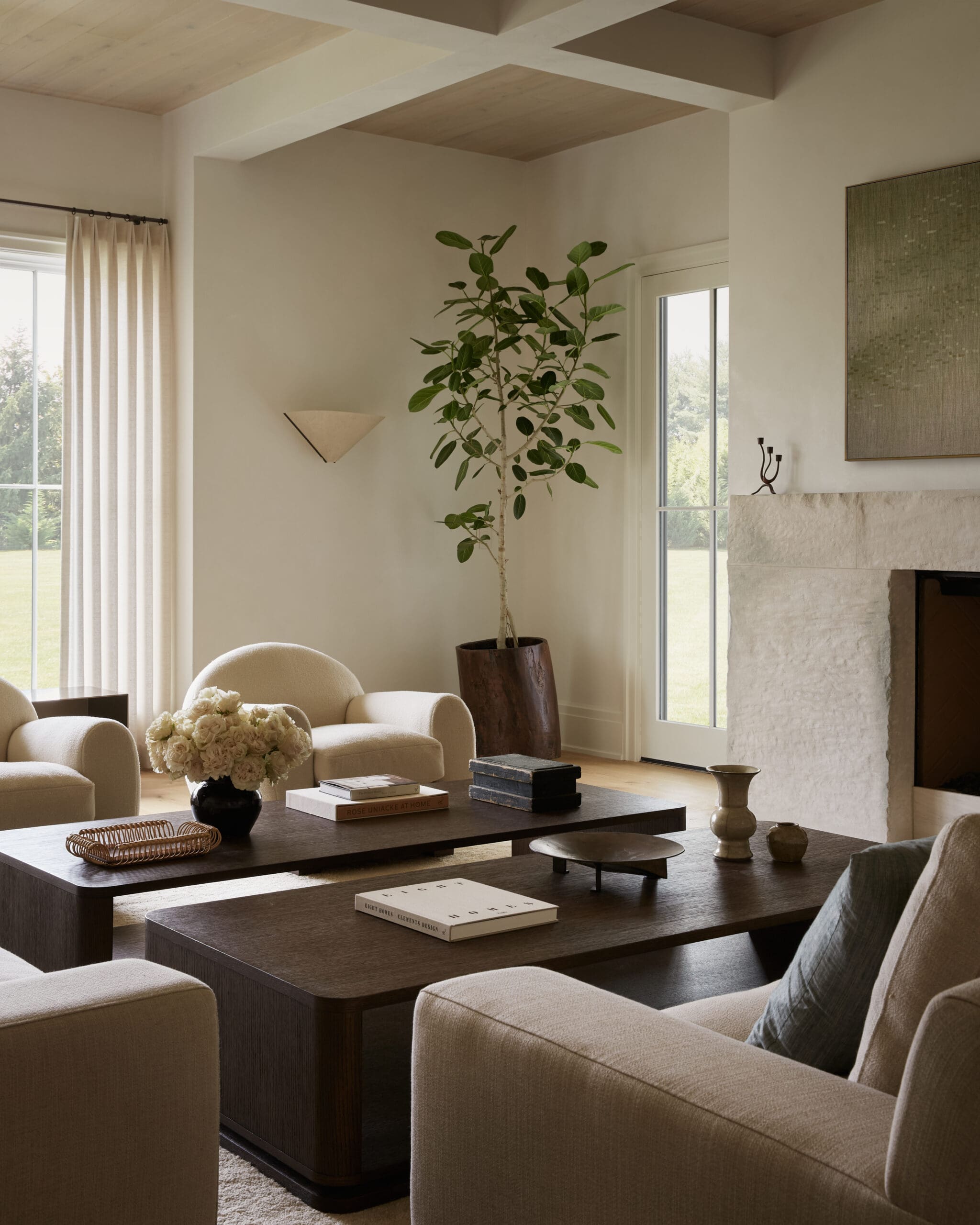 Living room with beige upholstered seating around a dark wood coffee table, a tall potted plant near a window, and a stone fireplace.
