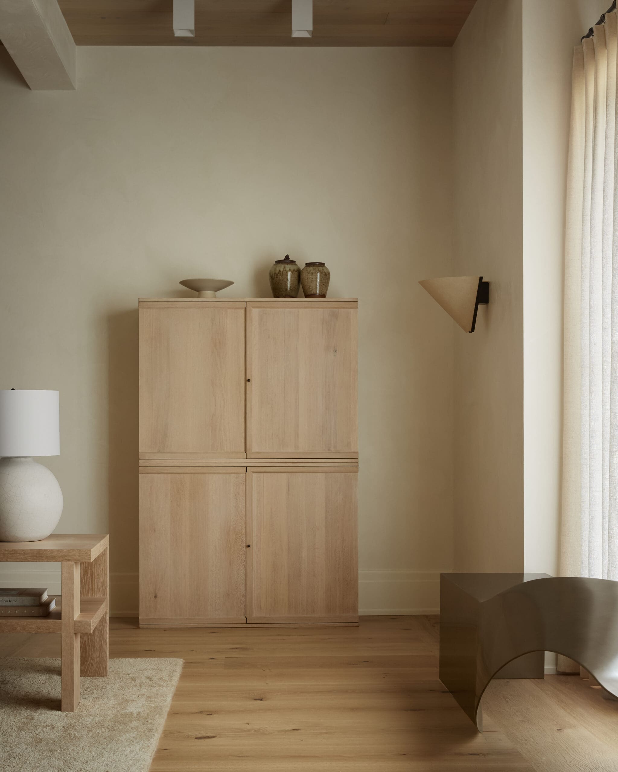 Minimalist living room corner with light wooden cabinet, two pottery jars, and a shallow bowl on top; beige walls and wood floor extend to a curtain on the right.