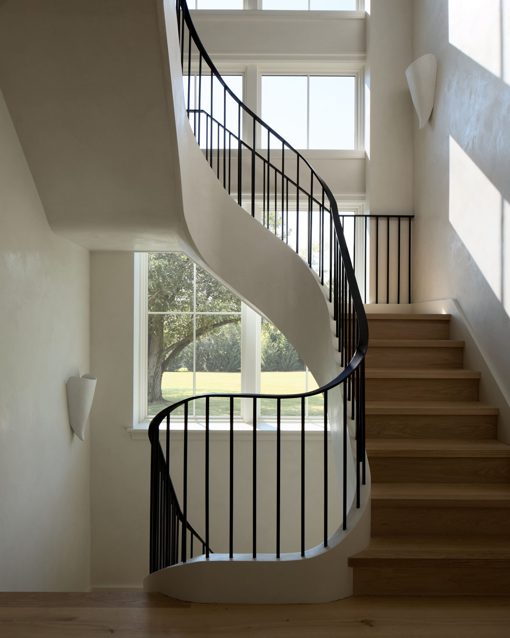 Curved white staircase with black metal railing and large daylight-filled windows showing a lawn outside, in a modern home.