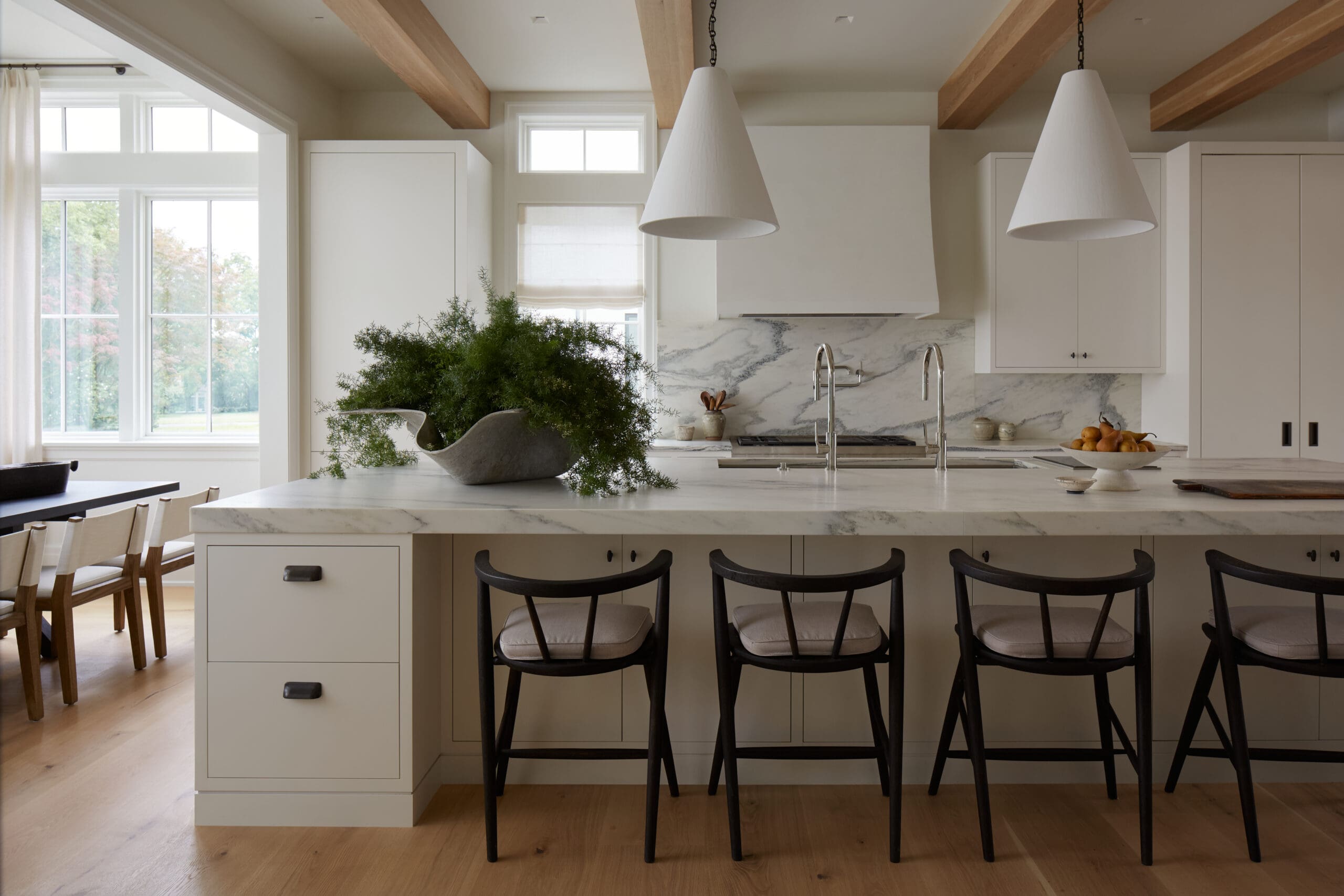Bright kitchen with a marble island, white cabinets, and two cone pendant lights; exposed wooden ceiling beams and natural light from windows.