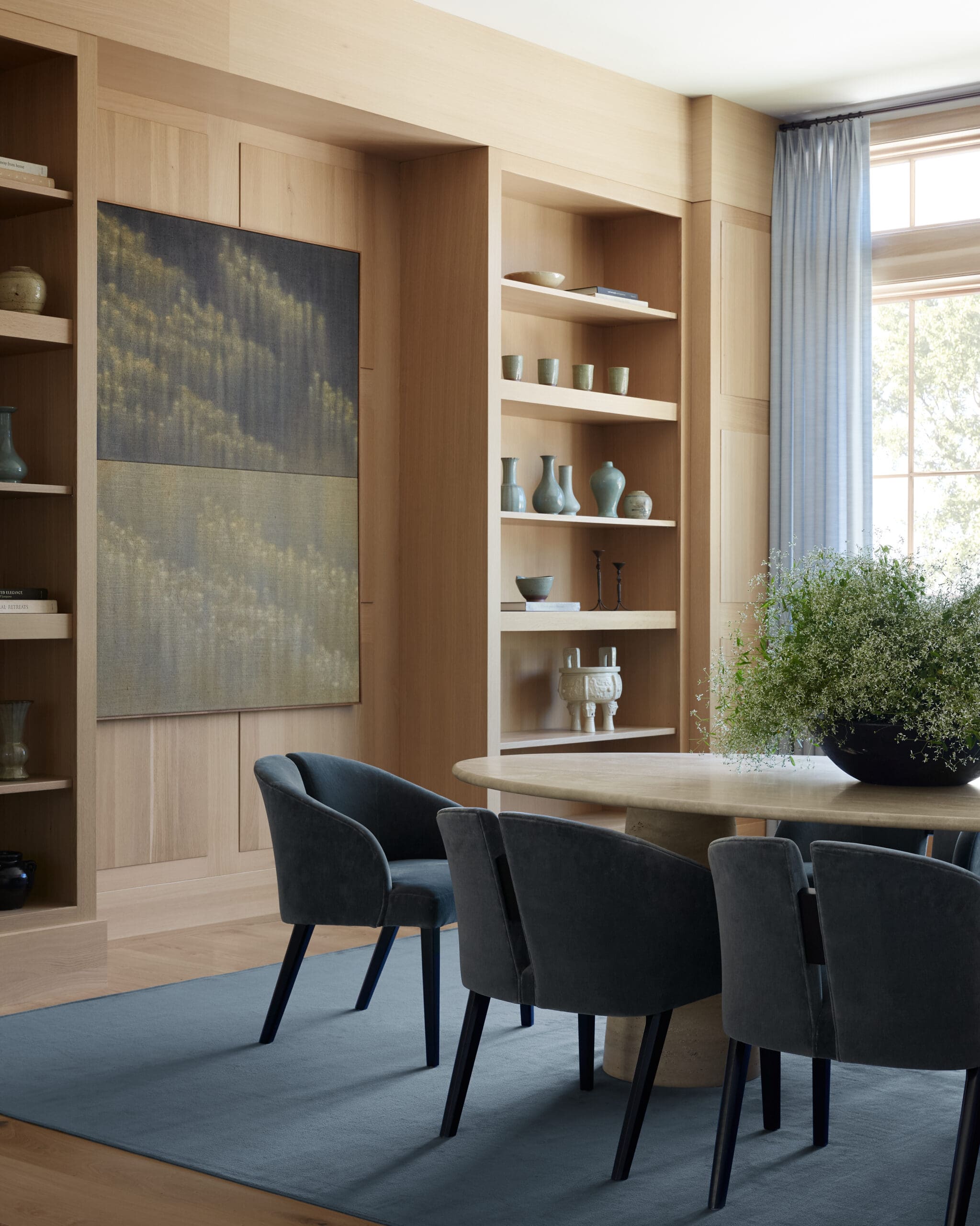 Round wooden table with blue upholstered chairs in a bright room with tall built-in shelves and a large window with blue curtains.