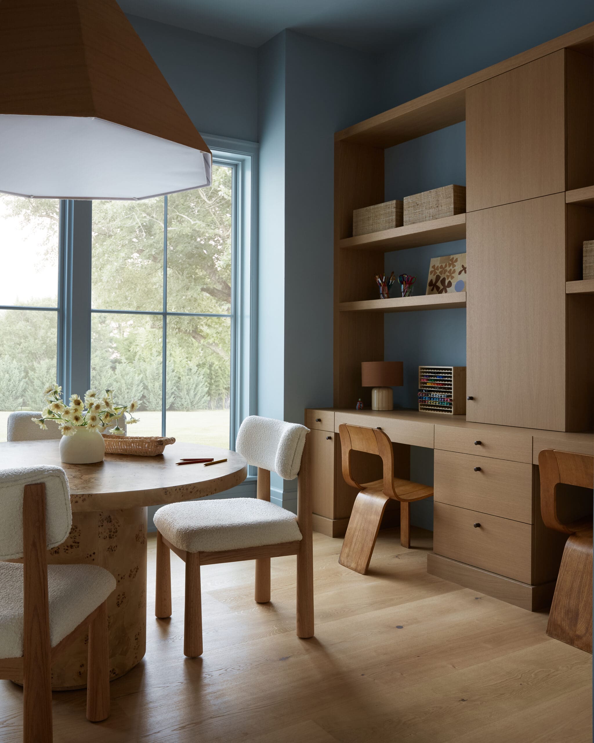 Round wooden dining table with beige upholstered chairs by a large window, and tall light-wood shelving unit against blue walls.