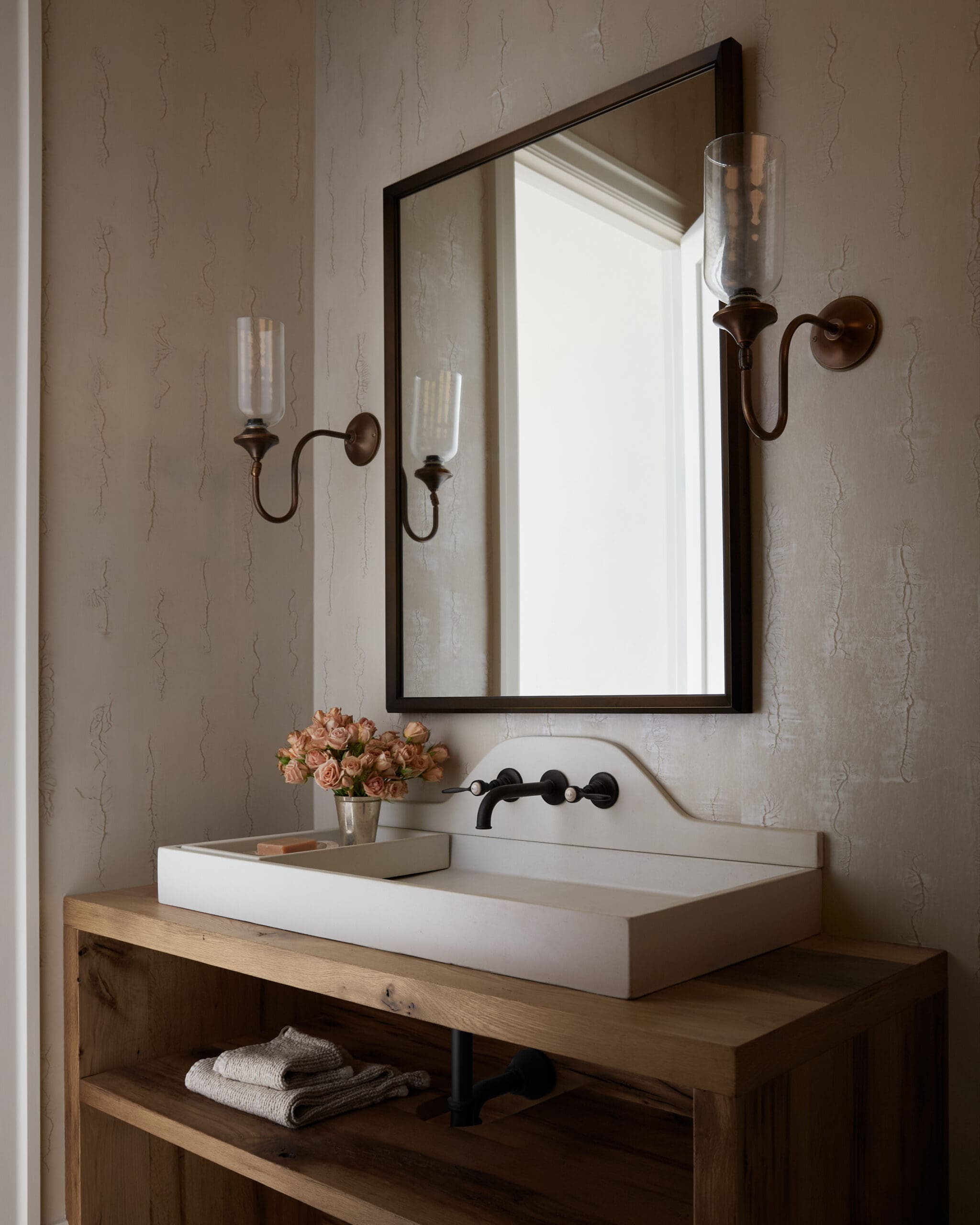 Rectangular white sink on a wooden vanity with a black faucet, framed mirror, bronze wall sconces, and a vase of pink roses on the counter.