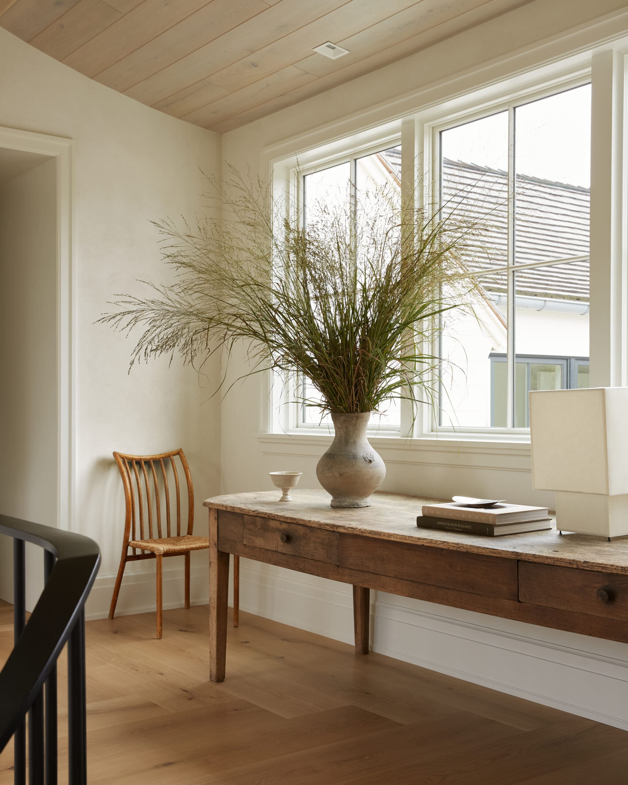 A rustic wooden desk with a large ceramic vase of tall dried grasses by a window, in a bright neutral room.