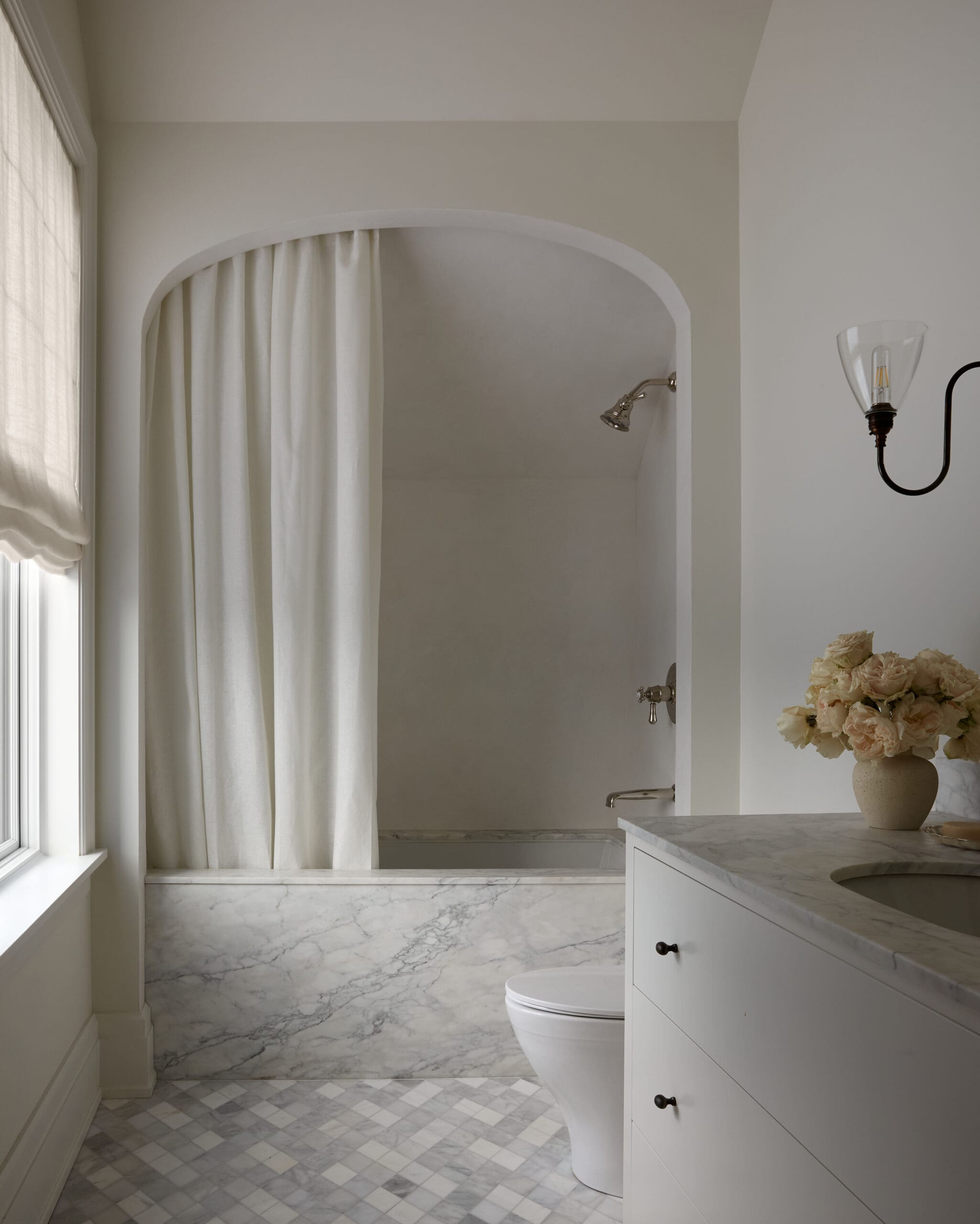 Bright white bathroom with marble vanity, built-in tub and curved shower opening, white curtains, and a pale rose bouquet in a vase.
