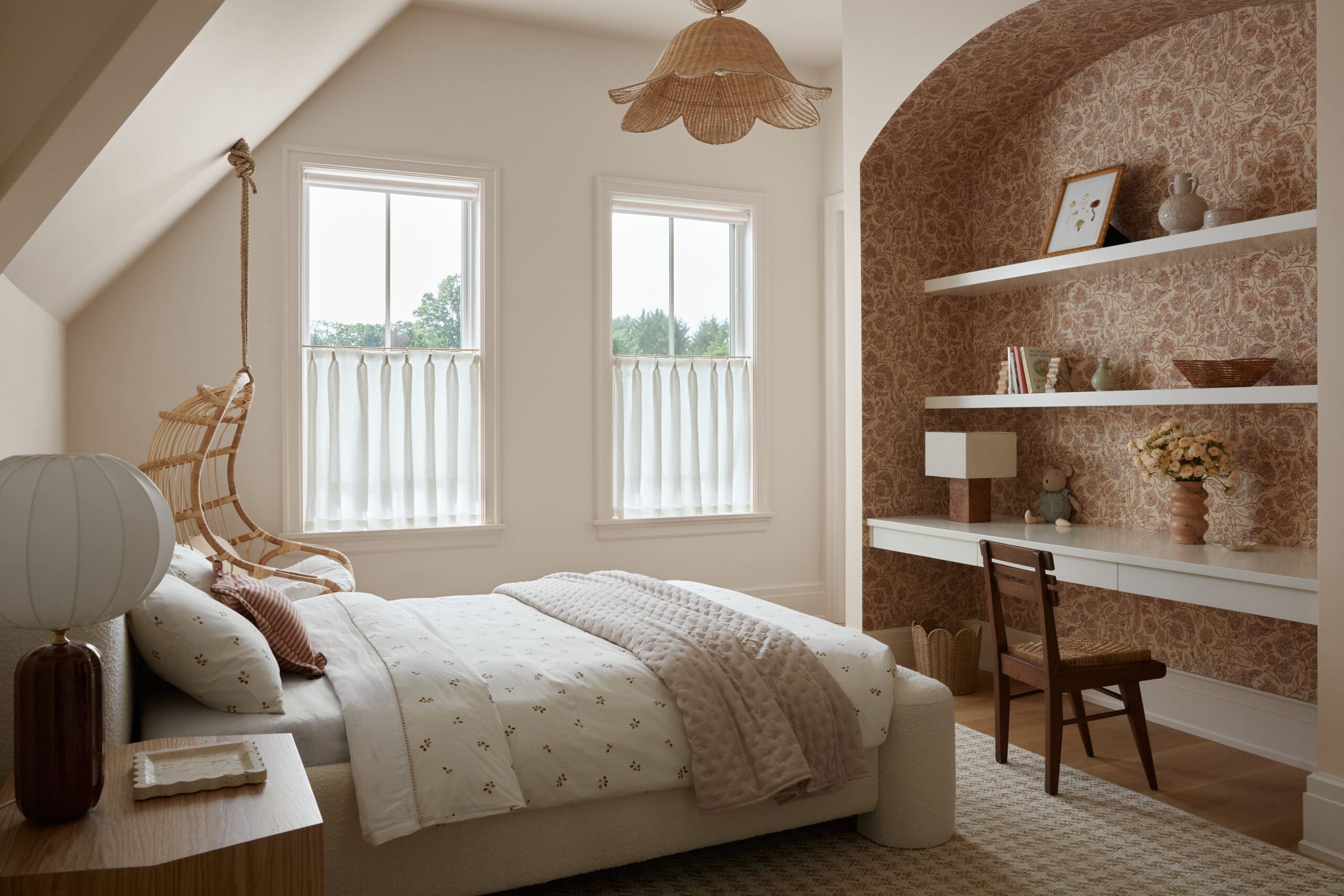 Cozy bedroom with two windows, sheer curtains, and a beige tufted bed; wicker chair and warm wallpapered wall on the right side