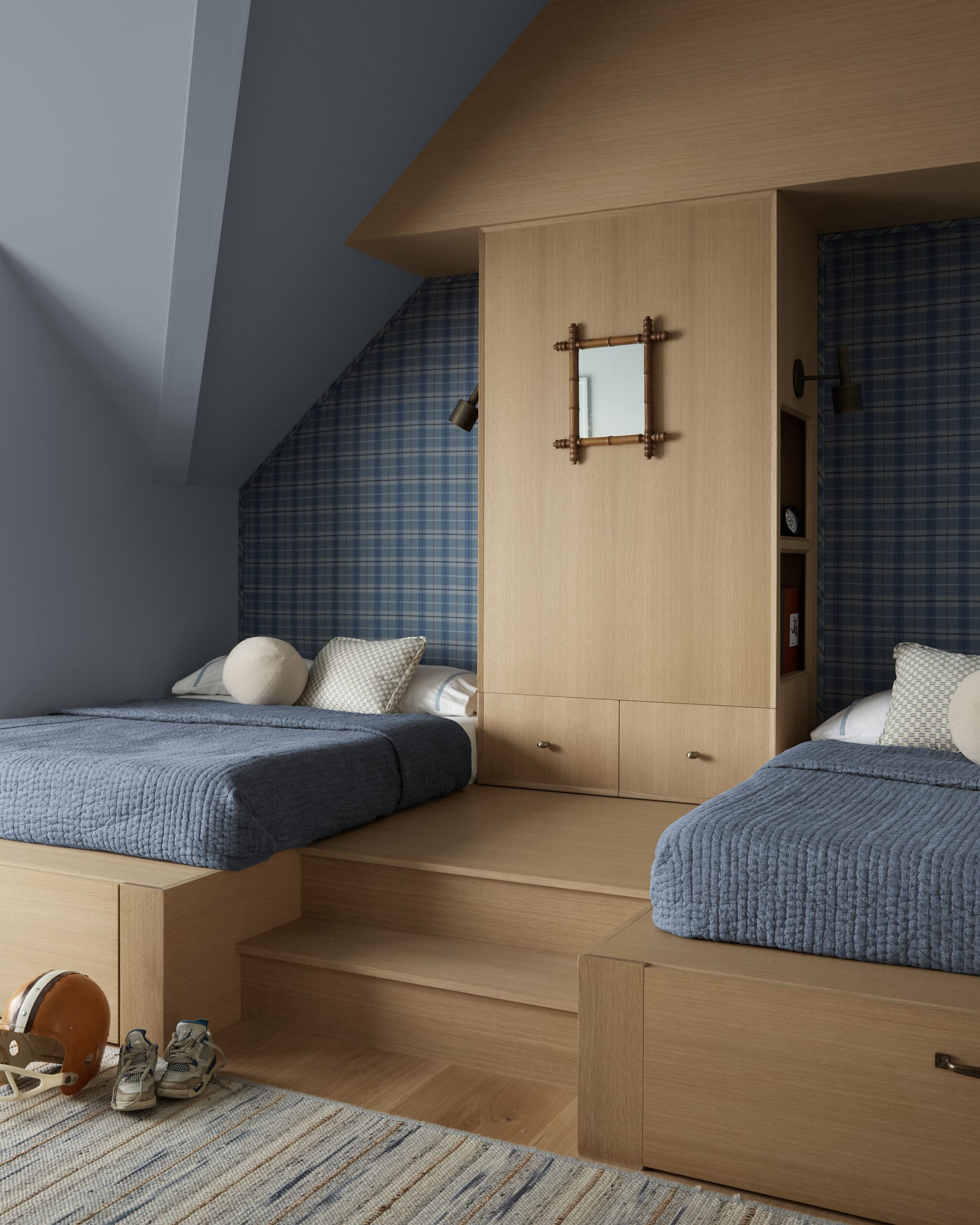 Two twin beds with blue quilted covers set in a light wood built‑in unit, with plaid blue wallpaper and a few shoes and a helmet on the floor nearby.