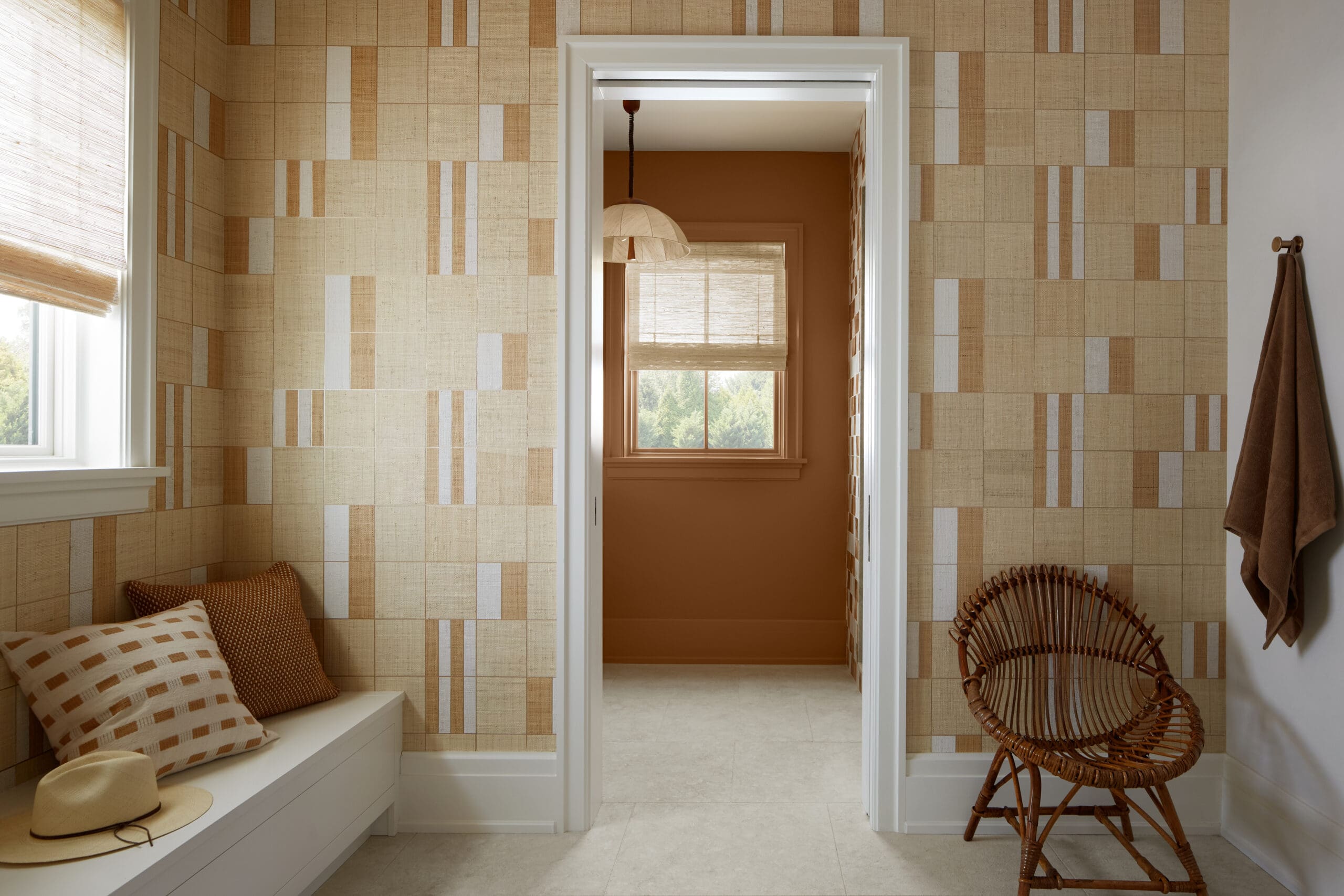 Doorway leads from a beige patterned-walls room into a warm brown-toned dressing space with a wicker chair and a straw hat on a white bench, plus brown cushions nearby.