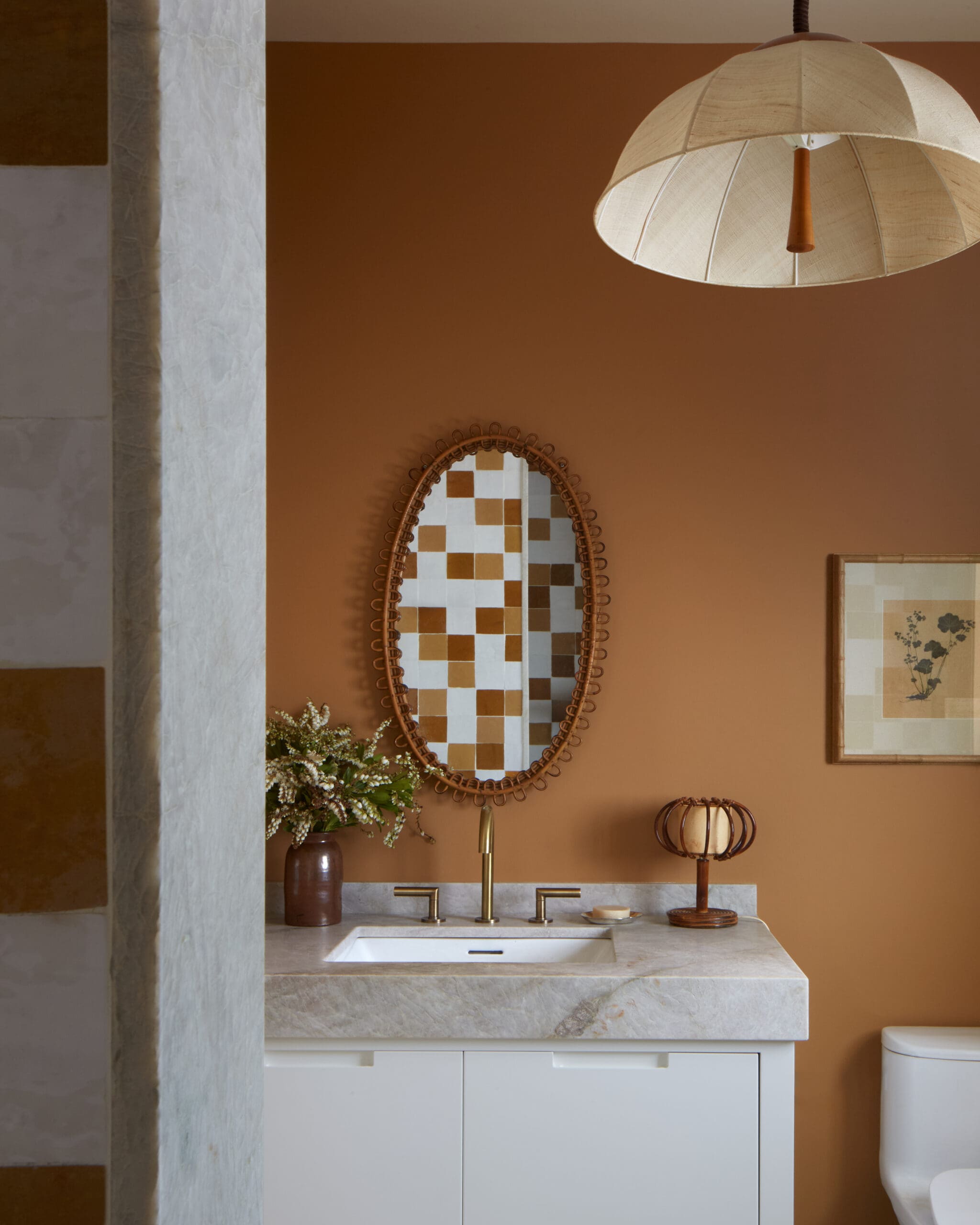 Bathroom vanity with white cabinets, marble countertop, and gold faucet set against a warm brown wall with a wicker oval mirror above."
