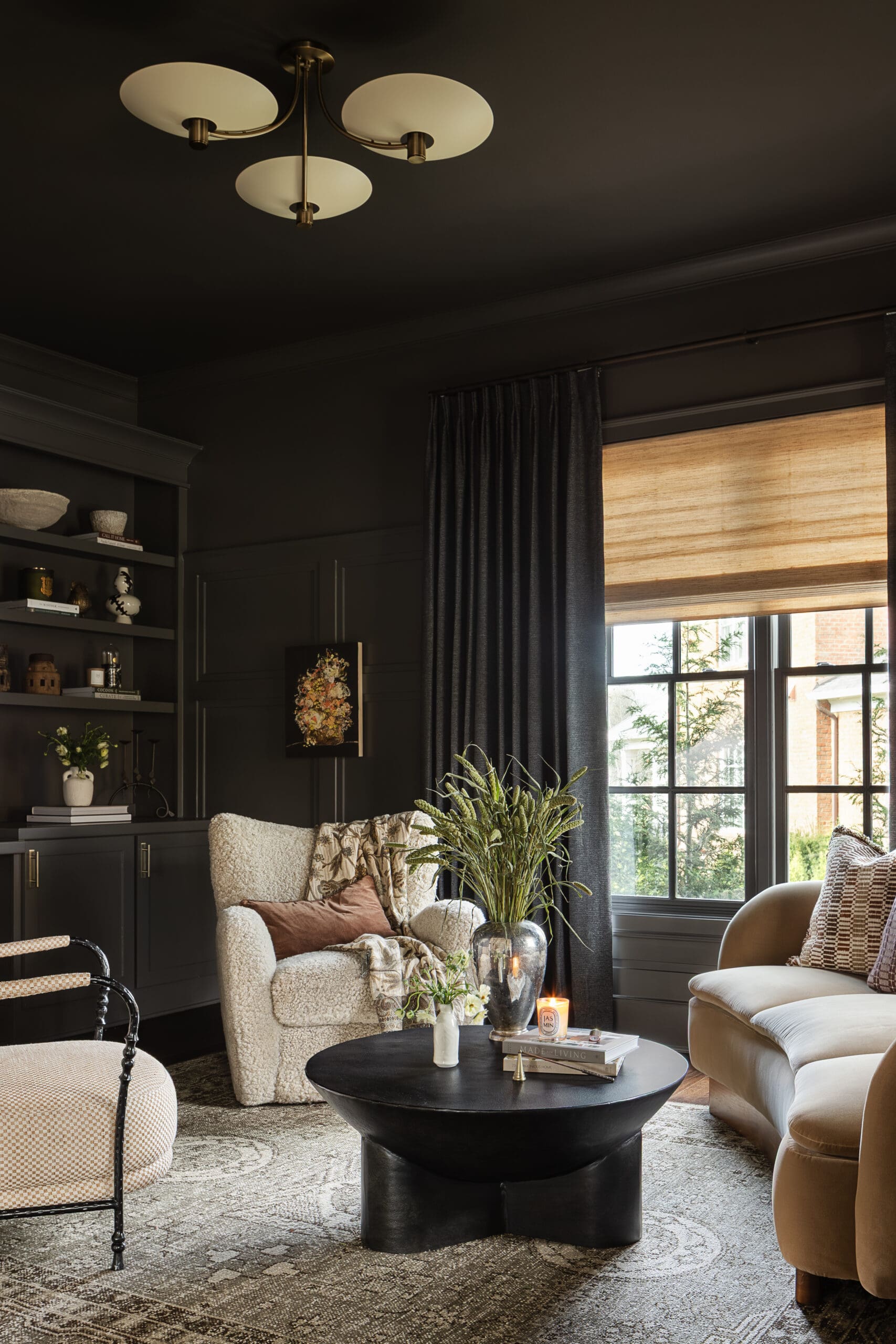 Cozy living room with dark walls, a black coffee table, and a beige sofa; large window with blinds and dark curtains, and a bookshelf on the left.