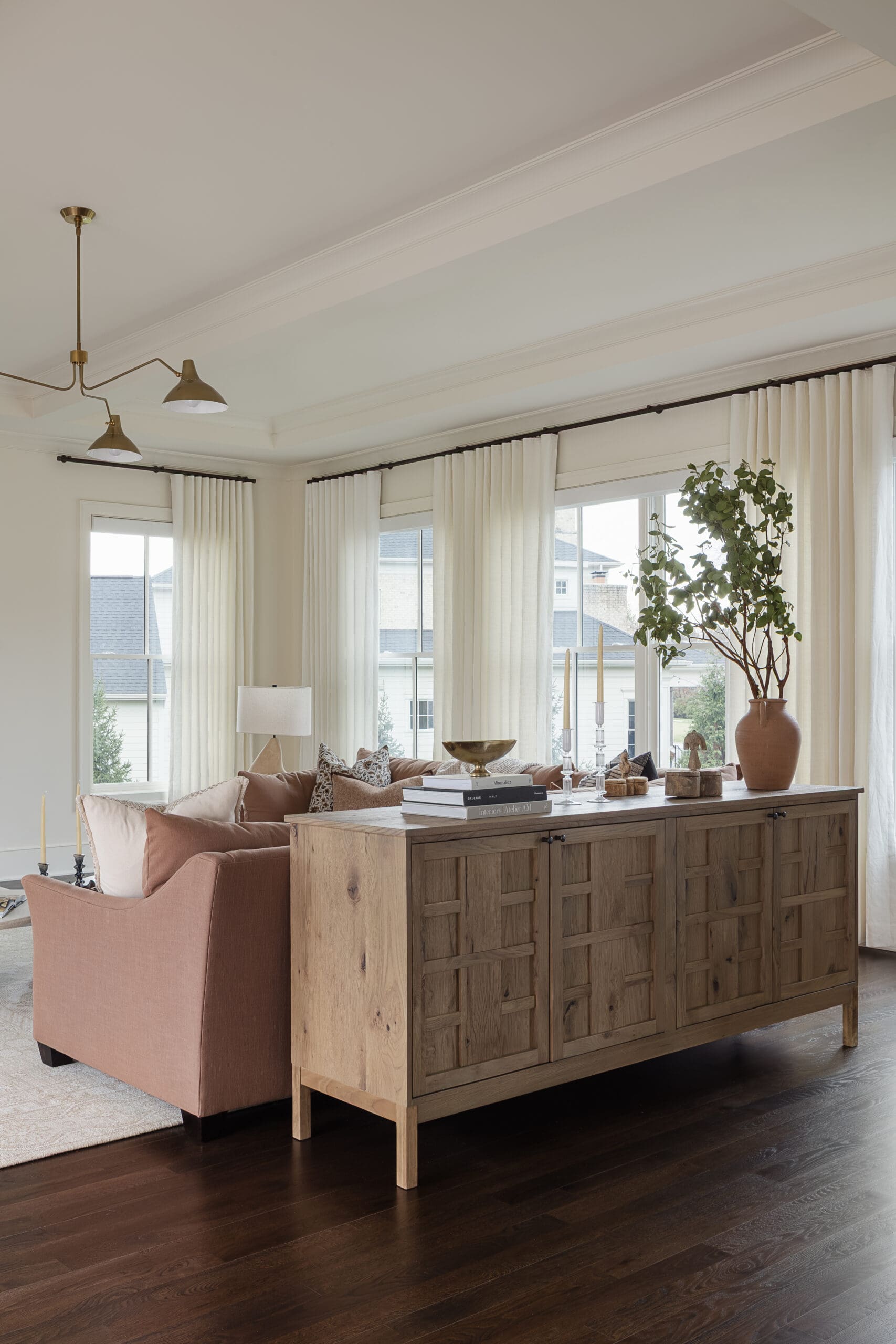 Sunlit living room with a carved wooden sideboard, pink sofa, white curtains, and a tall potted plant by the window.