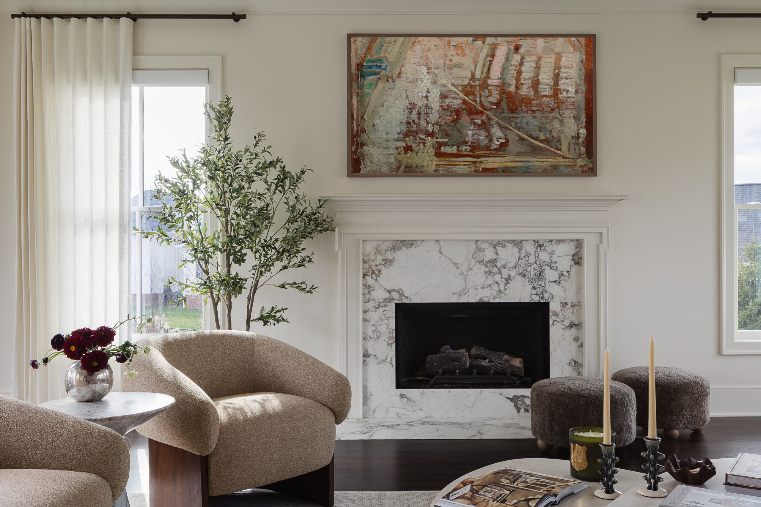 Living room with a marble fireplace, abstract wall art, and beige curved chairs; a tall plant by the window and a vase of flowers on a side table.