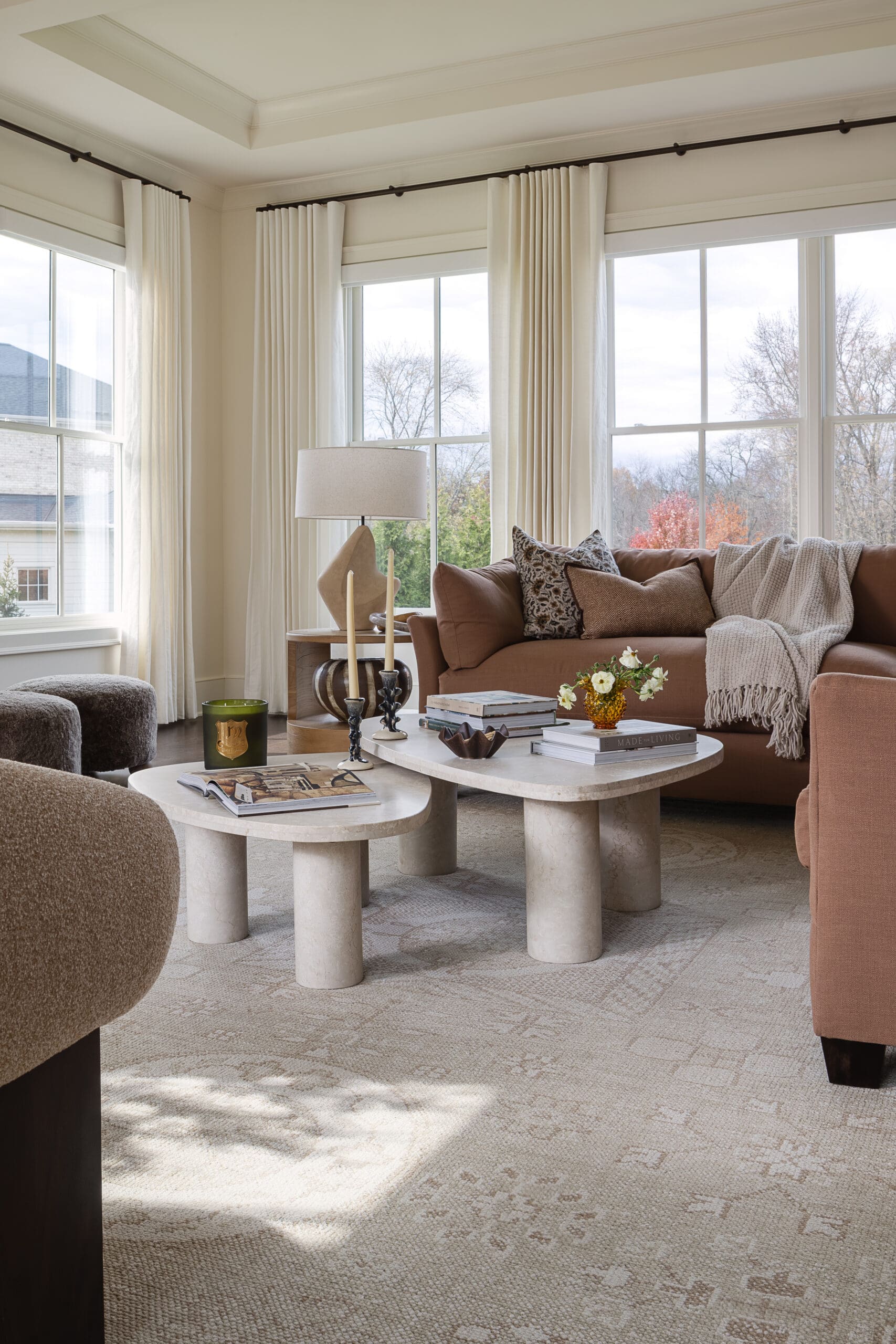 Living room with beige carpet, tan sofa, decorative pillows, and round stone coffee tables beneath large windows with white curtains. Natural light fills the space.