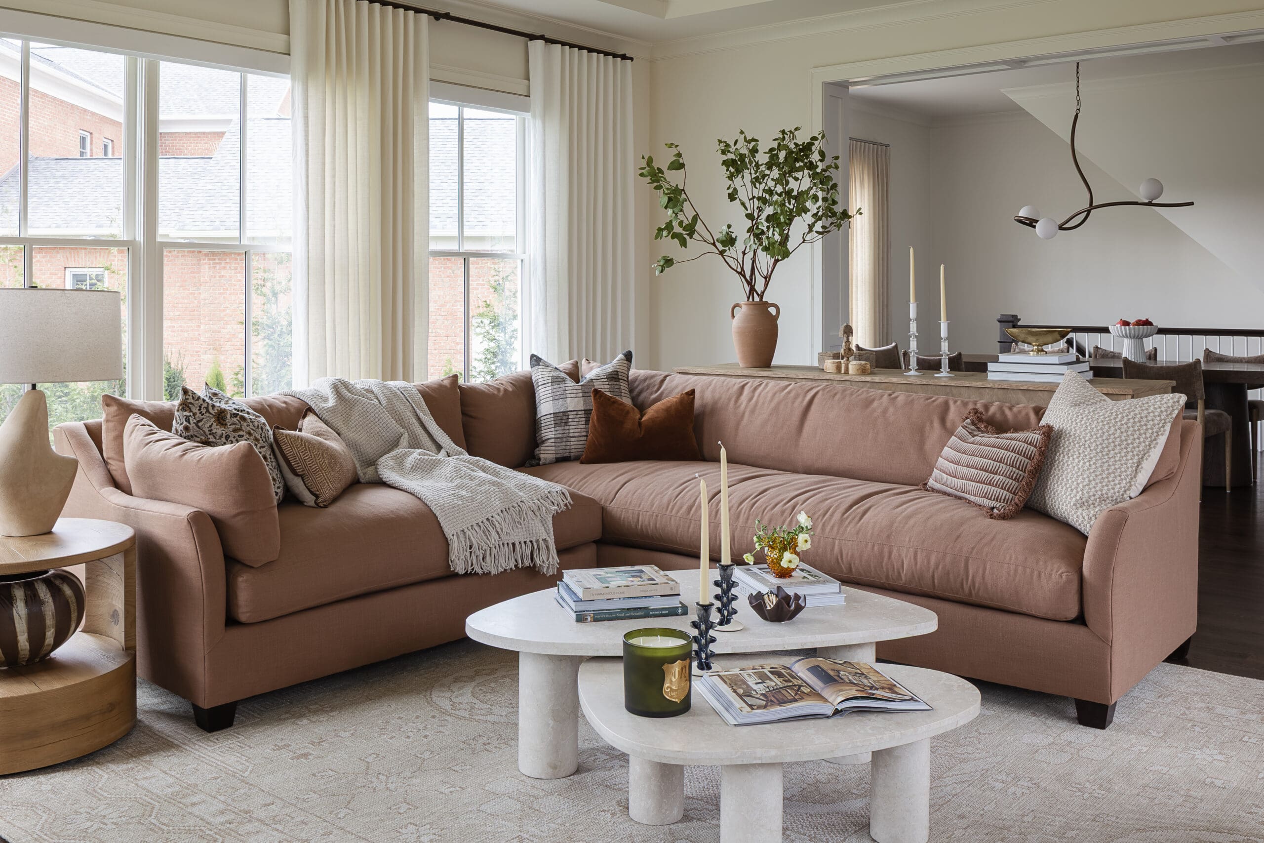 Cozy modern living room with a pink sectional sofa, pillows, and white marble coffee tables near large windows.