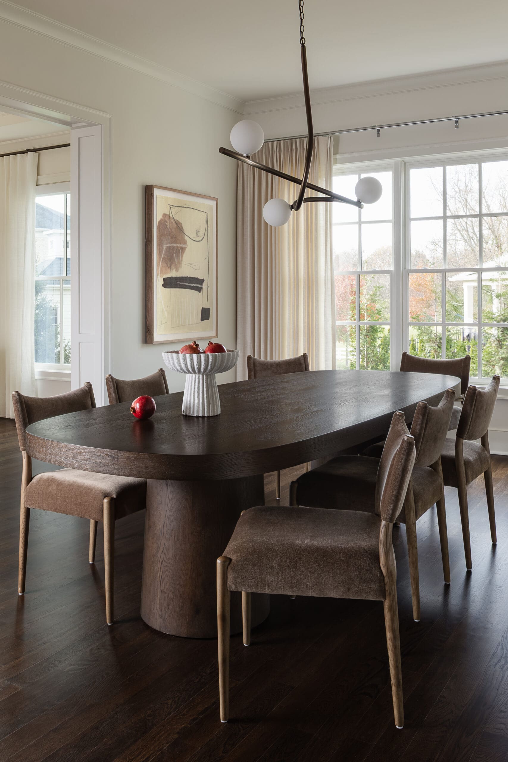 Dark oval dining table surrounded by brown upholstered chairs in a bright breakfast room with a white bowl of apples as a centerpiece and large arched windows behind.
