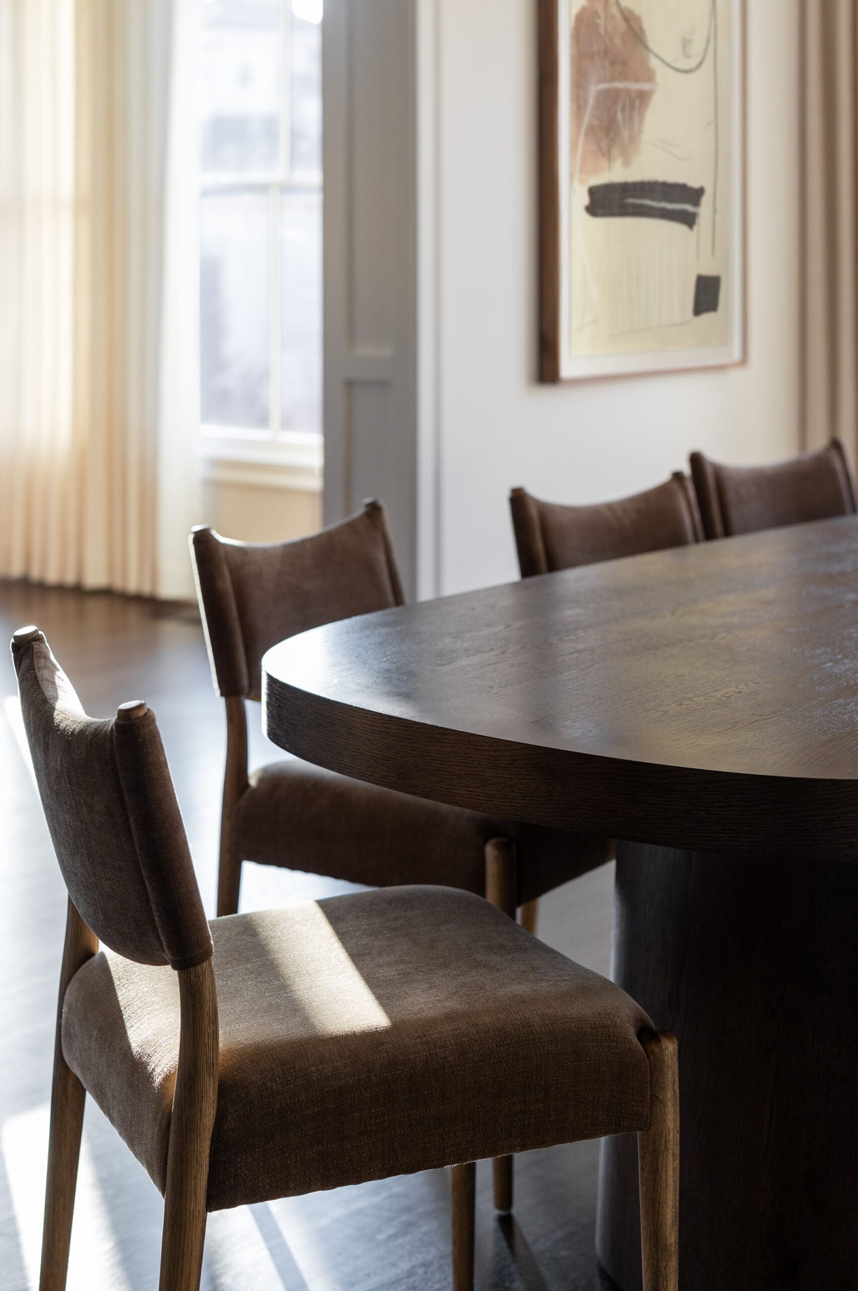 Round wooden dining table surrounded by brown upholstered chairs in a sunlit dining room with sheer curtains and a framed abstract painting on the wall