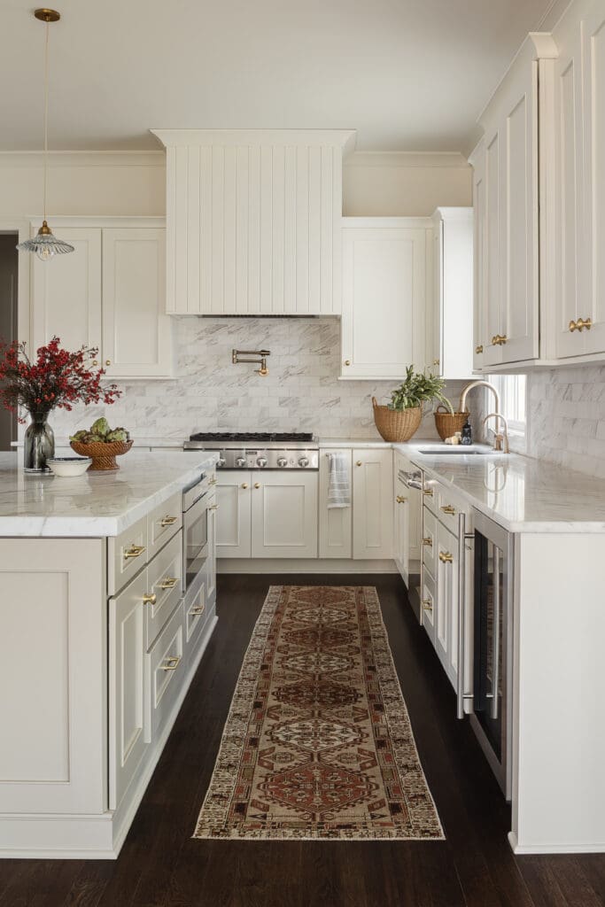 Bright white kitchen with marble backsplash, brass hardware, and a long patterned runner rug along dark wood flooring.
