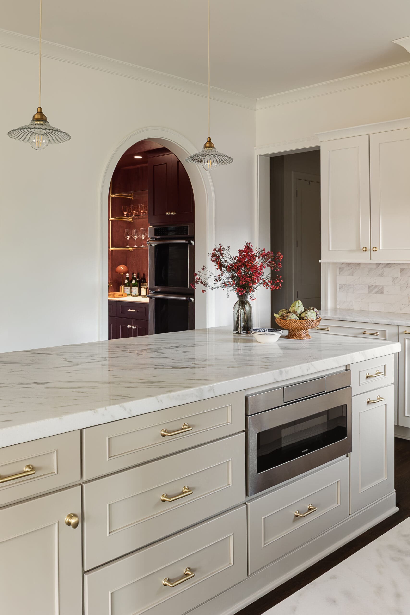 White kitchen with marble island, brass hardware, and a built-in stainless oven; burgundy arched doorway reveals a bar area.