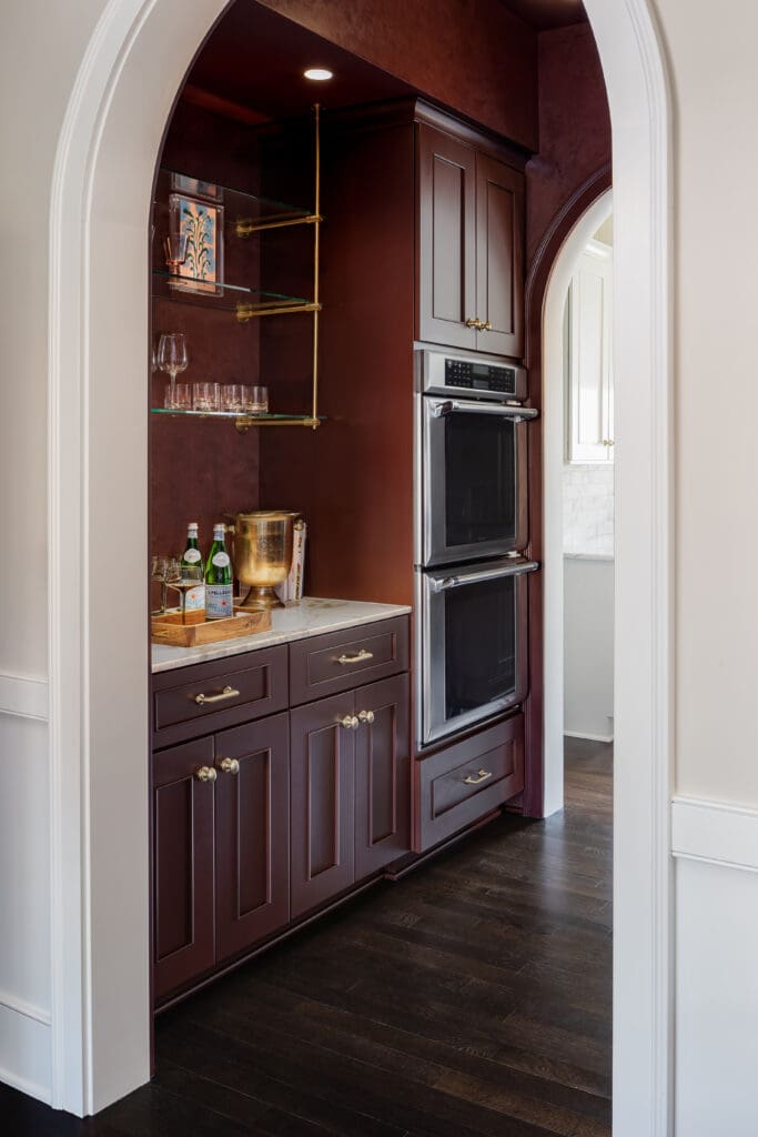 Built-in dark wood kitchen cabinetry with brass hardware, glass shelves, and a stainless double oven set in an arched doorway.