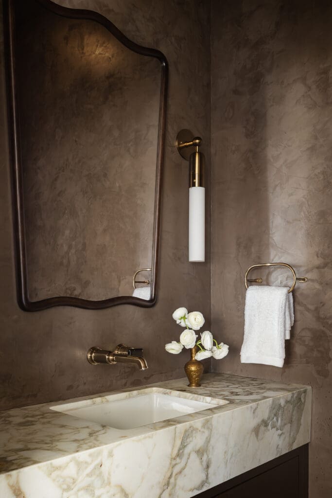 Elegant bathroom vanity featuring a curved bronze-framed mirror, brass faucet, and white towels on a marble countertop with a vase of white flowers nearby.