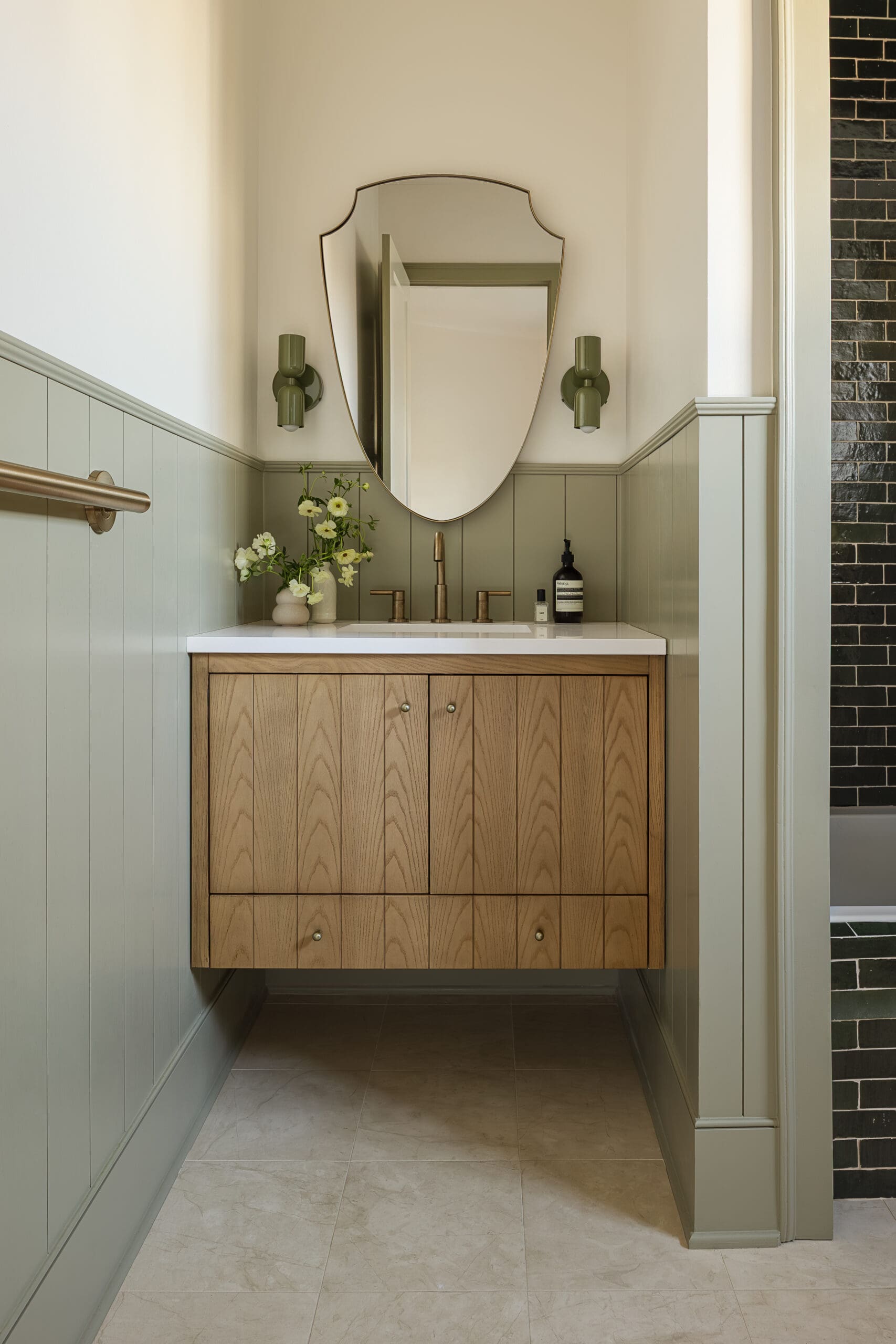 Wood vanity with white countertop and a shield-shaped mirror flanked by green wall sconces in a bathroom.